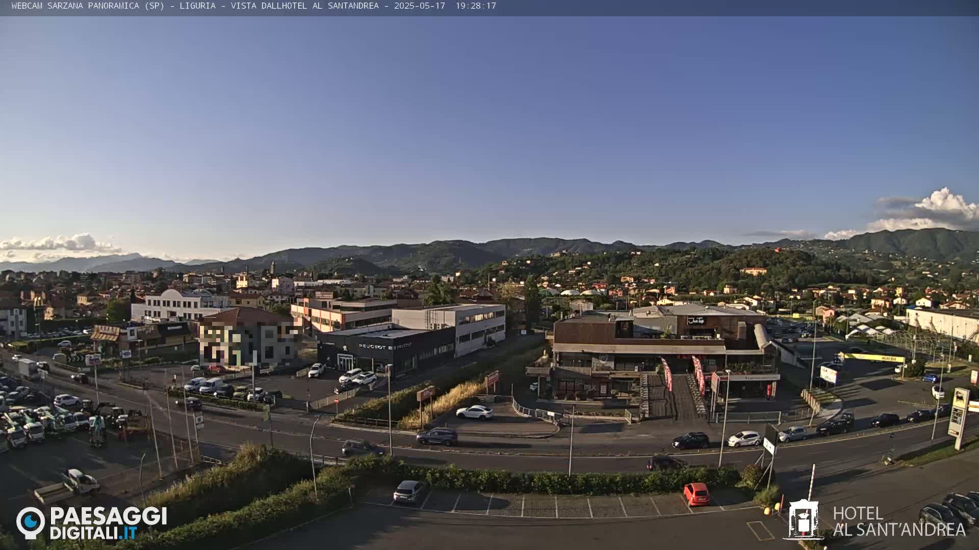 A clear, sunny day reveals a panoramic view of a town nestled in a valley surrounded by rolling hills, with a parking lot and various buildings visible in the foreground.