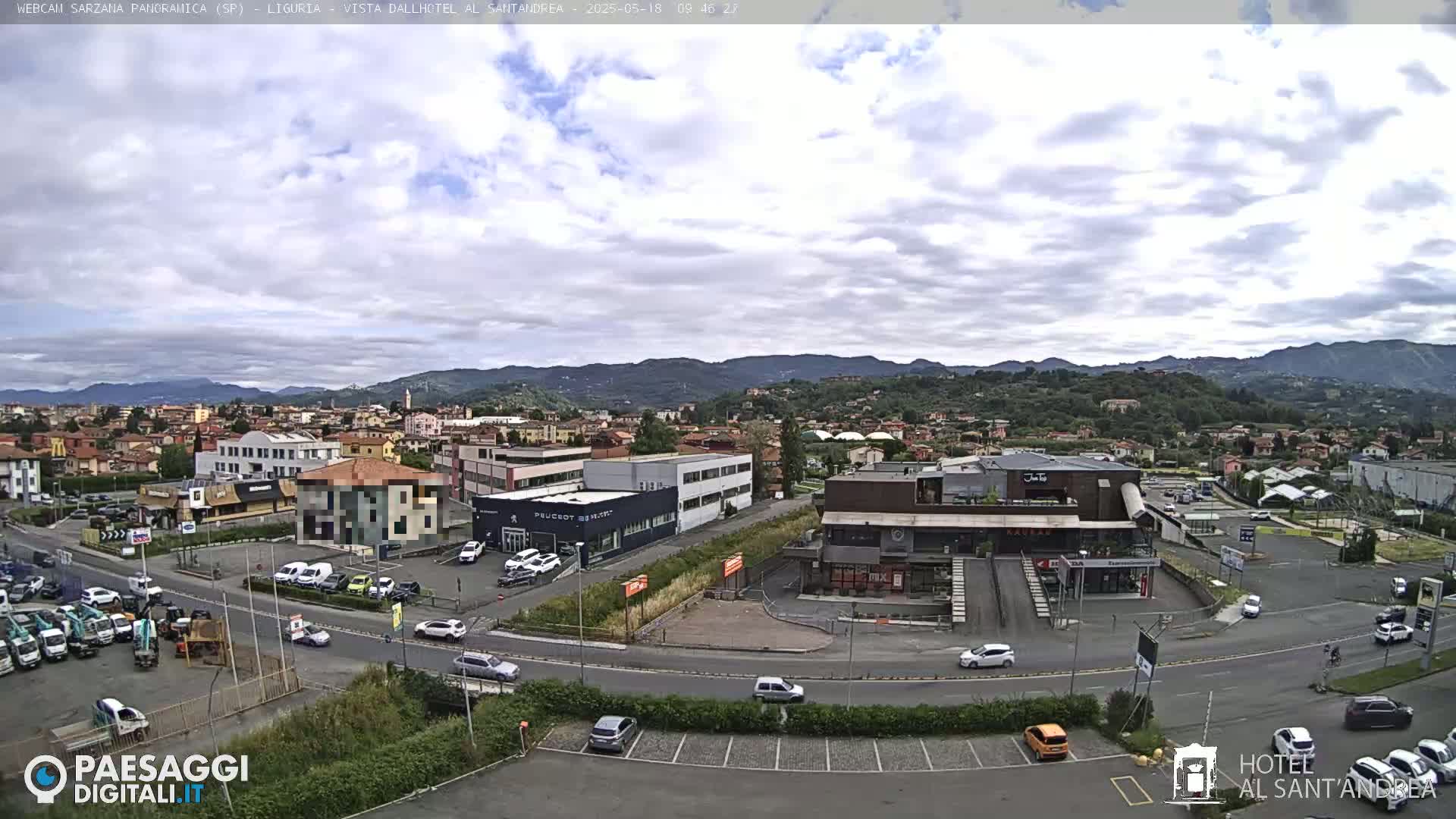 A partly cloudy day reveals a wide view of a town nestled in a valley between mountains, with a parking lot and several buildings in the foreground.