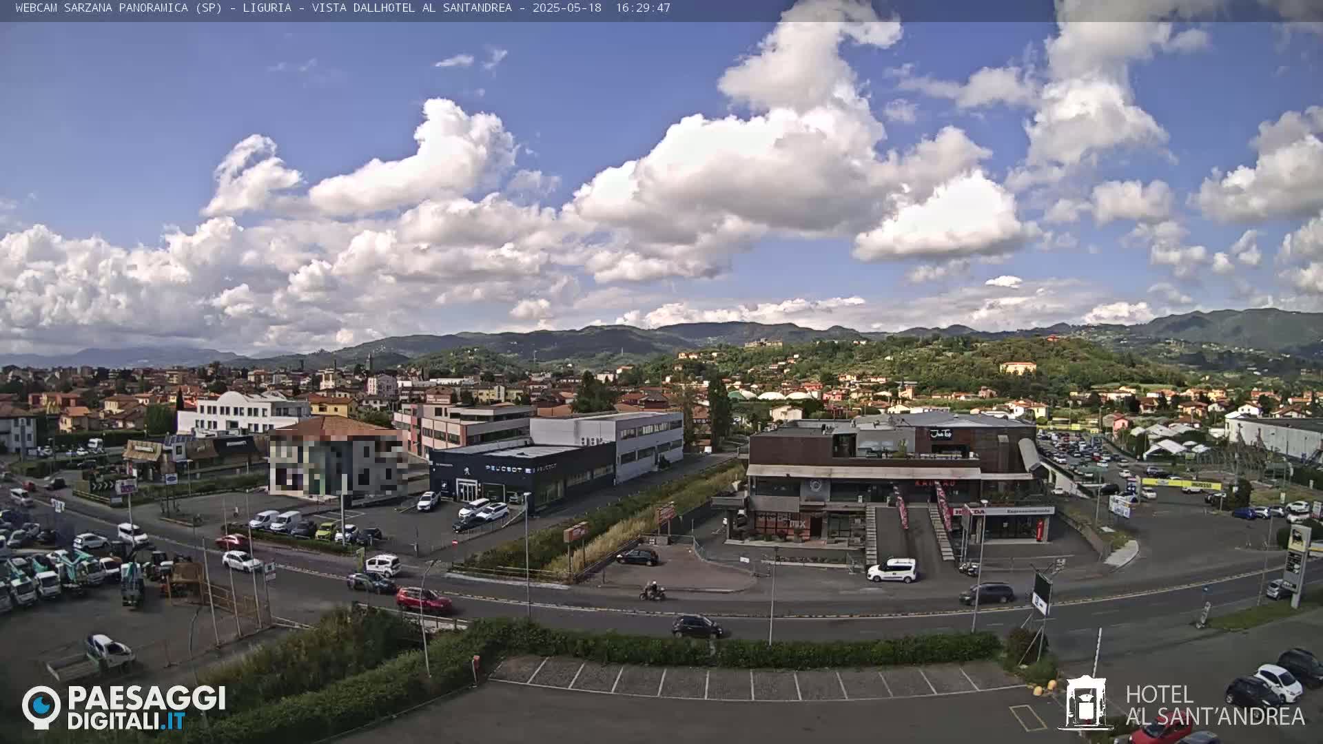 A partly cloudy day reveals a panoramic view of a town nestled in a valley, surrounded by green hills and mountains.