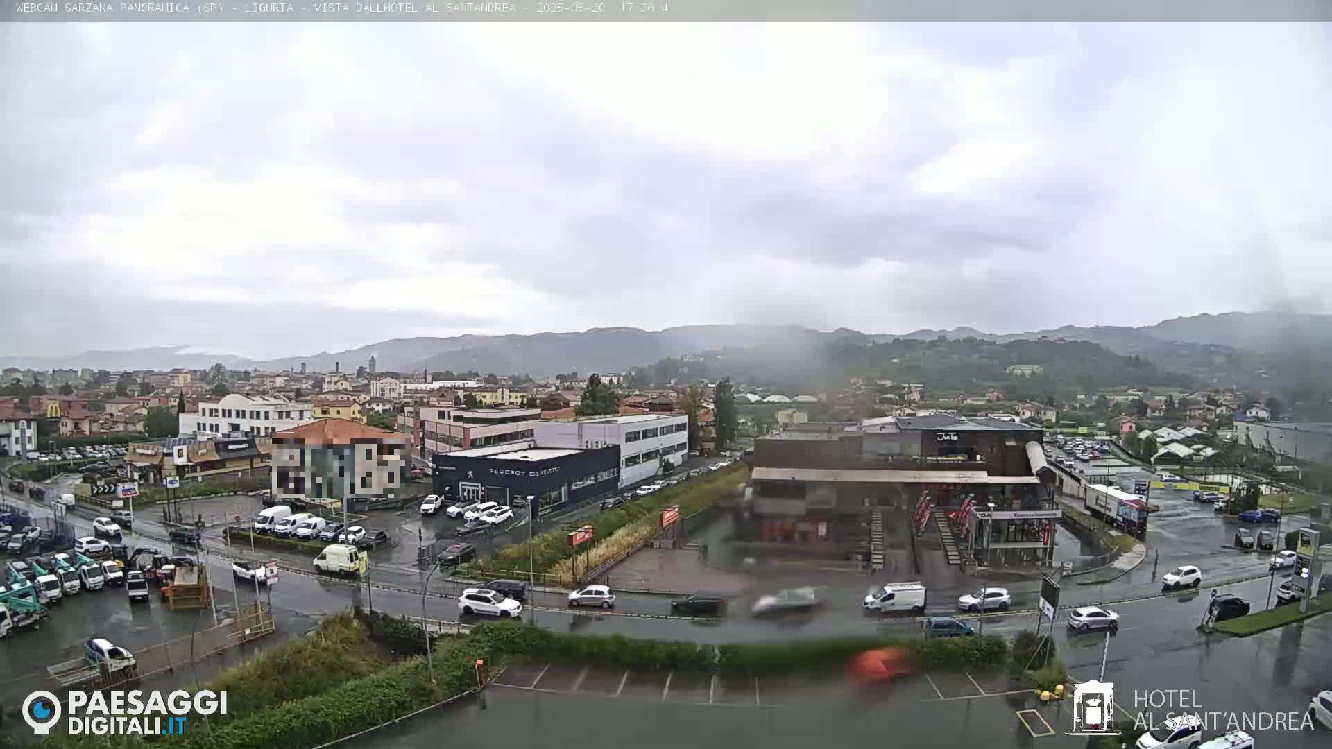 A rain-slicked town nestled in a valley between hills is viewed from an elevated position, showing cars traveling on wet roads and buildings scattered throughout.