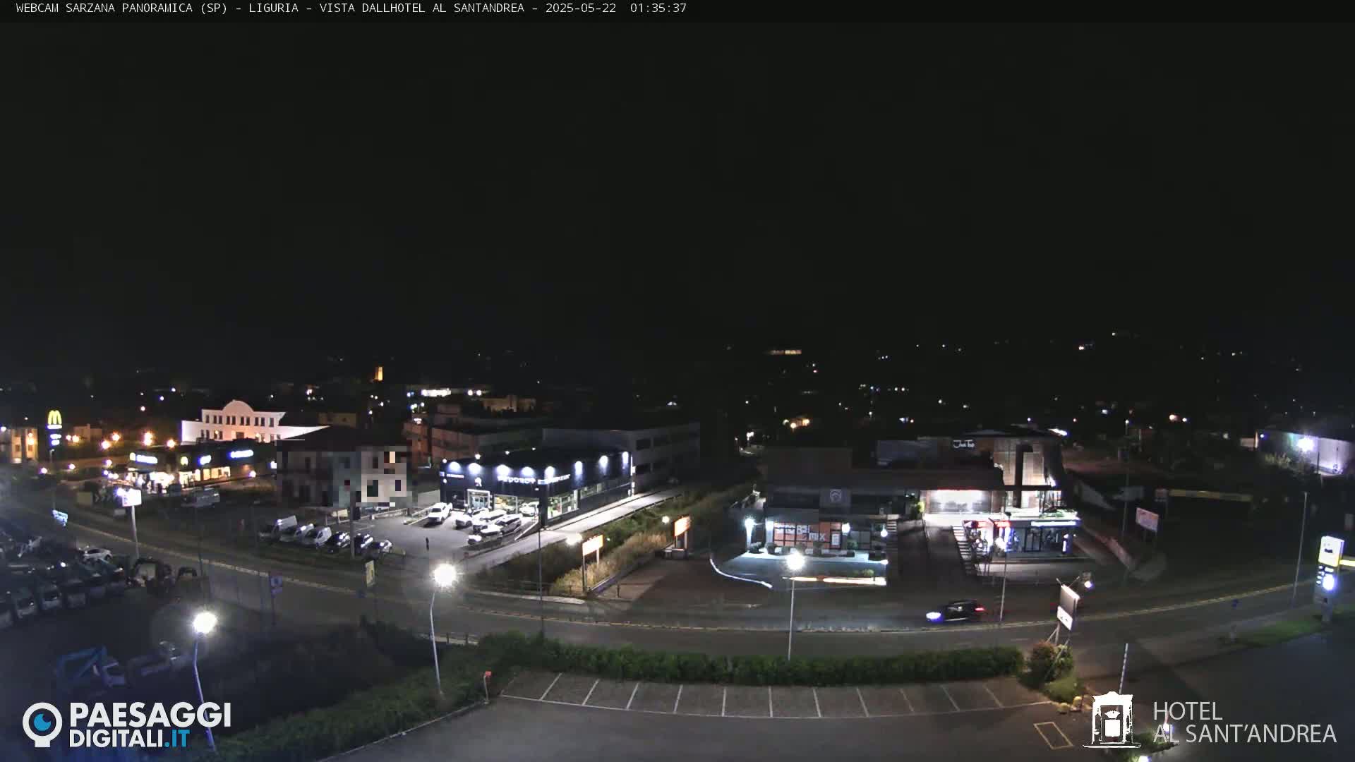 A nighttime, aerial view of a town's outskirts showing several buildings, roadways, and parked cars.