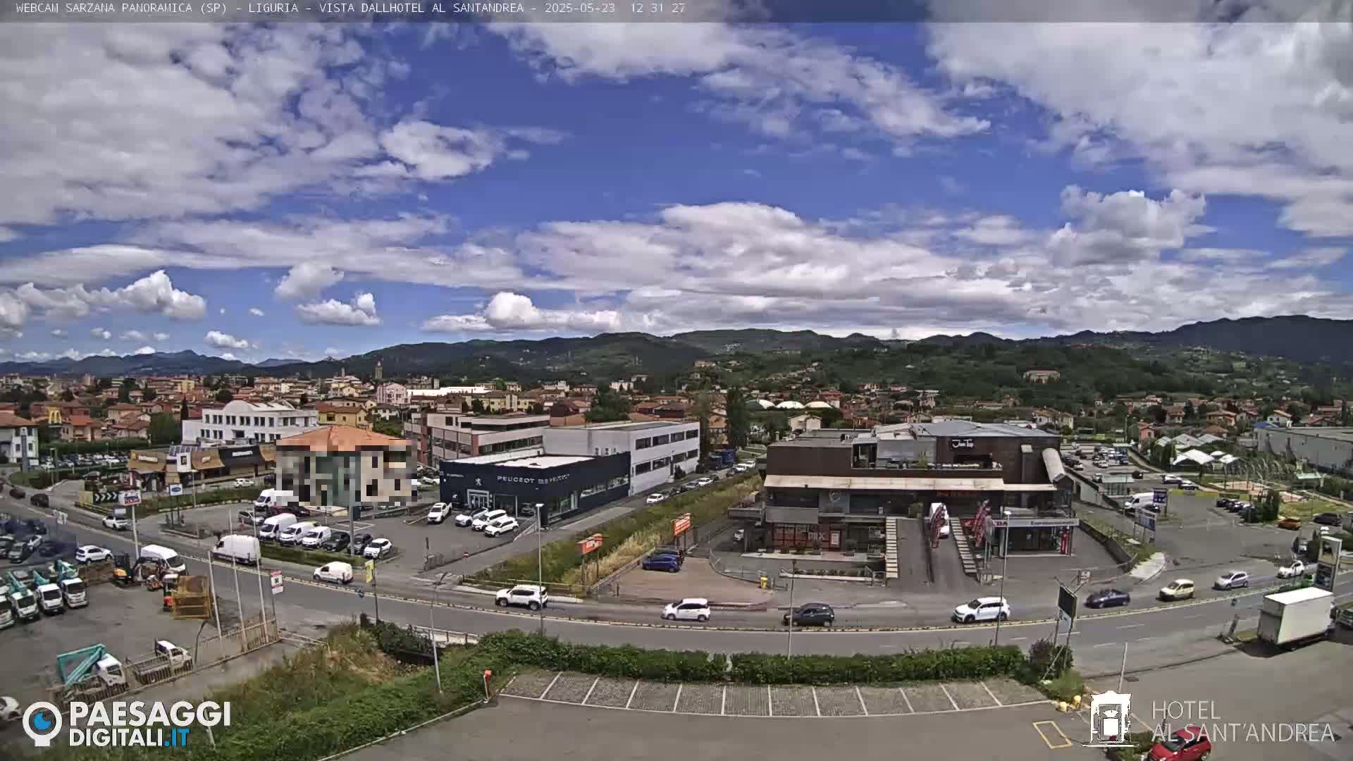 A partly cloudy day reveals a view of a town nestled amongst hills, with numerous buildings and cars visible in a wide shot.