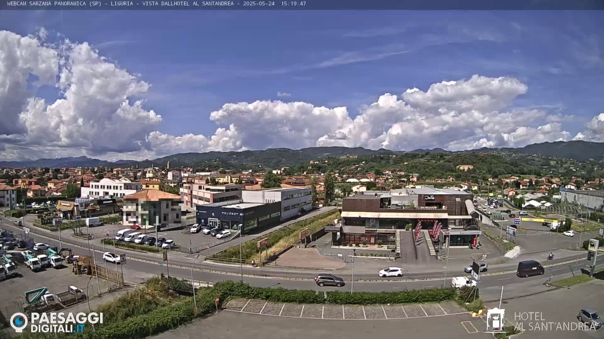 A partly cloudy day reveals a panoramic view of a town with buildings, roads, parked cars, and distant hills.