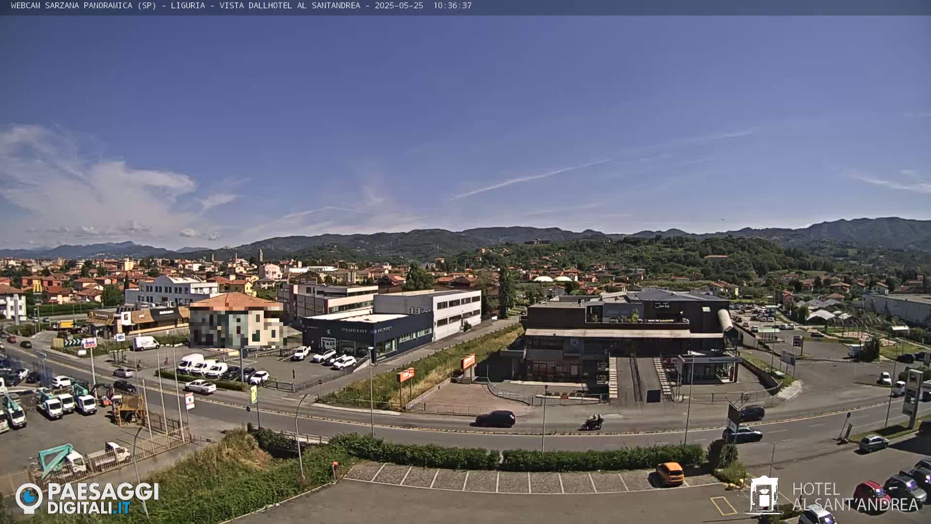 A sunny day reveals a sprawling town nestled in a valley, surrounded by hills and mountains, with cars parked in lots near several buildings.
