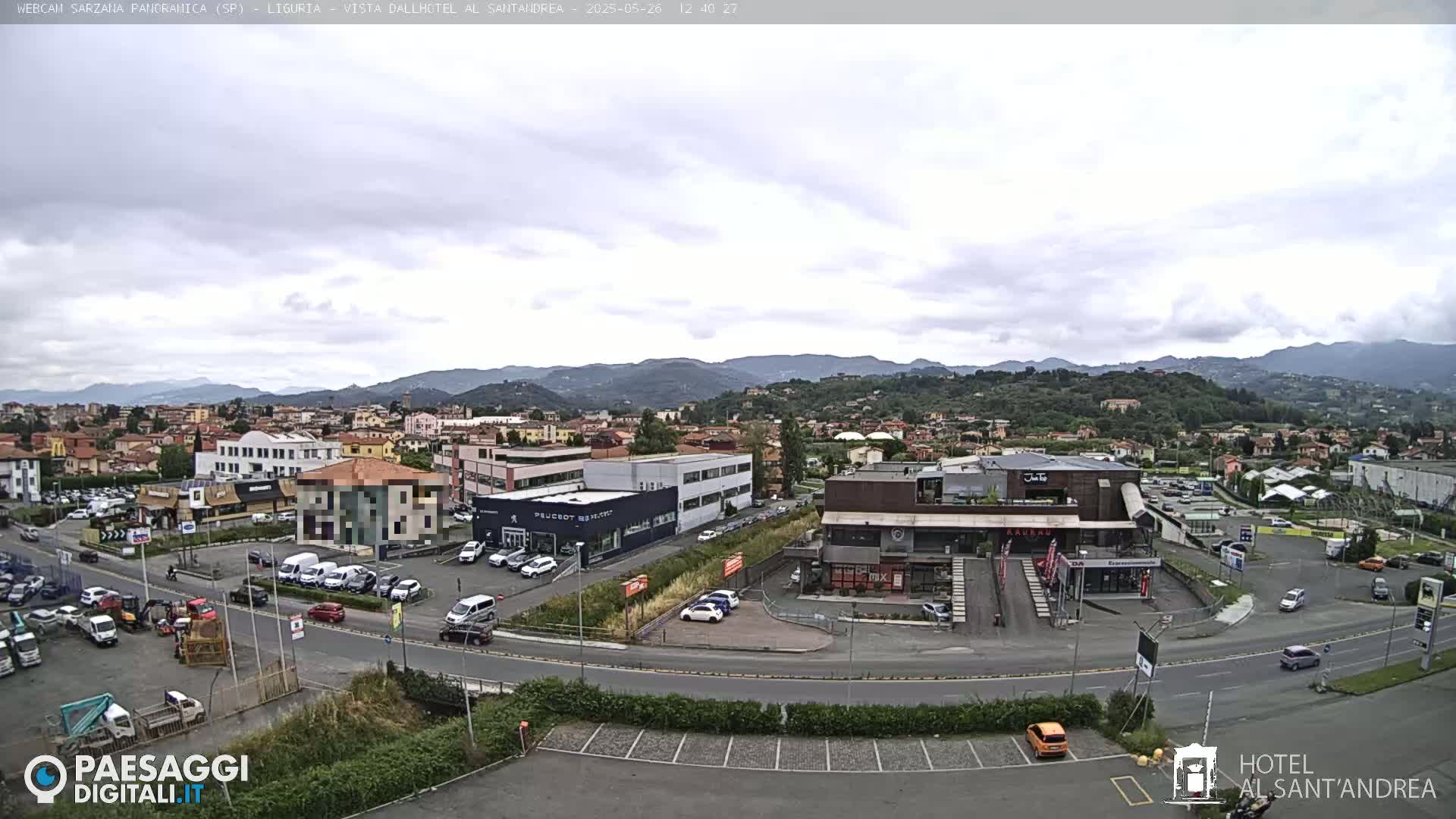 A partly cloudy day reveals a wide shot of a town nestled in a valley, surrounded by rolling hills, with various buildings, roads, and parked cars visible.