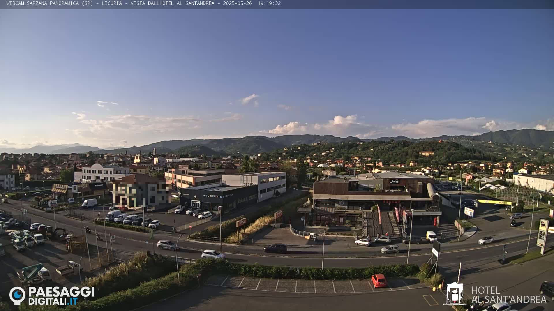 A partly cloudy afternoon view of a town nestled in a valley, with mountains in the background and cars parked in a large lot in the foreground.