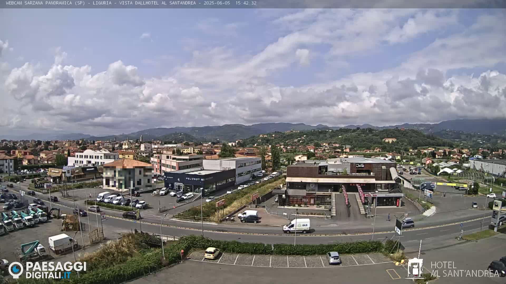A partly cloudy day reveals a panoramic view of a town nestled between hills, with cars parked in a lot near various buildings, including a car dealership and a restaurant.