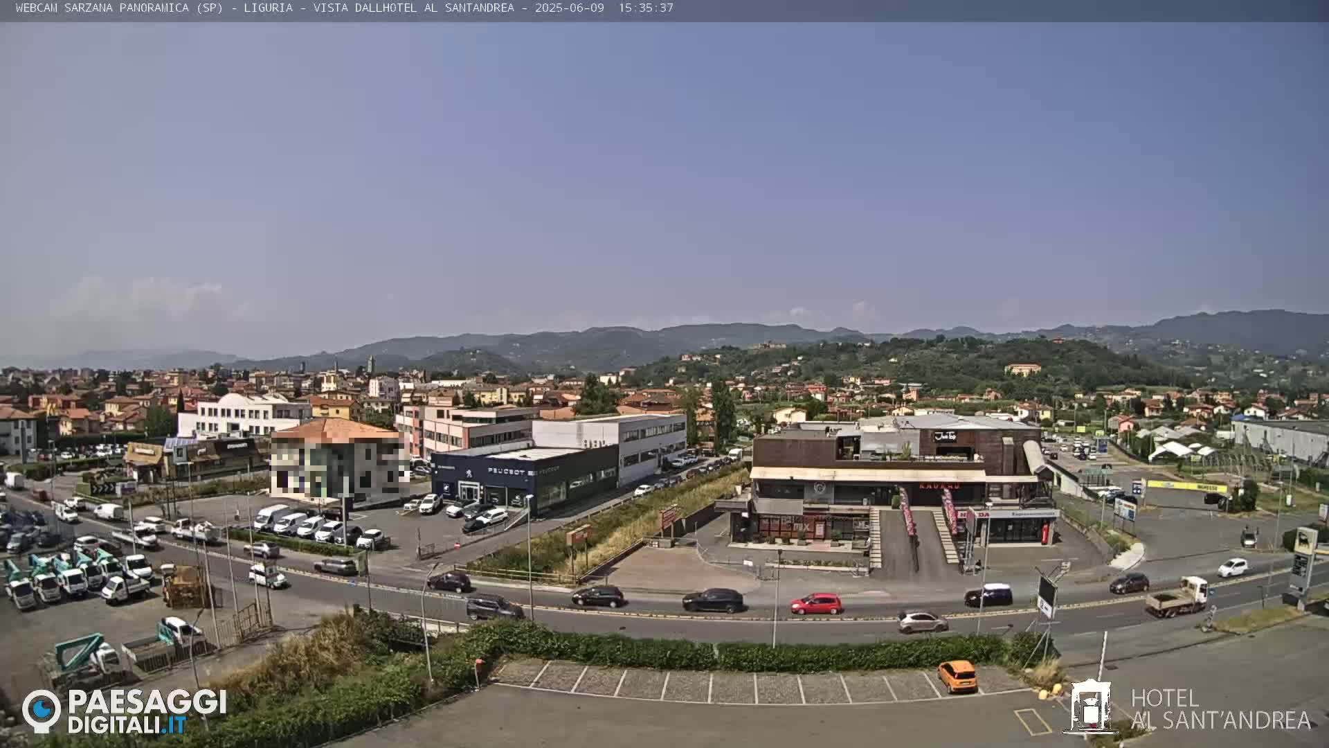 A sunny day reveals a panoramic view of a town nestled among rolling hills, with vehicles moving along roads and parking lots near various buildings.
