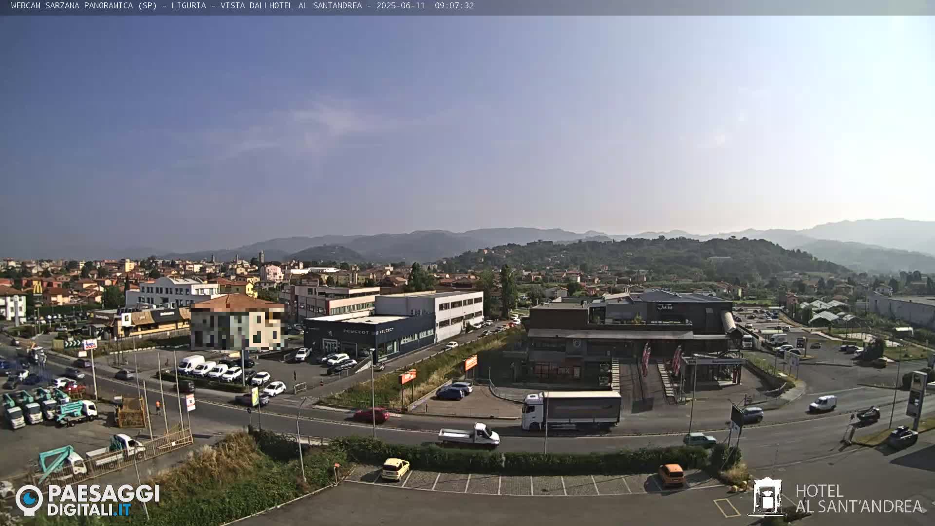 A sunny, clear day reveals a panoramic view of a town nestled in a valley, surrounded by distant mountains, with numerous cars and buildings visible, including a hotel and a car dealership.