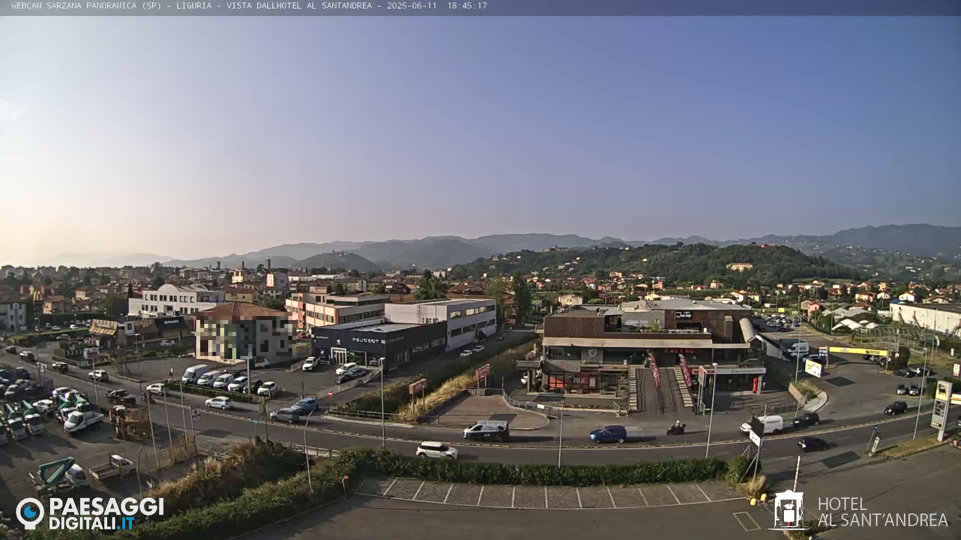 A clear, sunny day reveals a panoramic view of a town nestled among rolling hills, with various buildings, cars, and a parking lot visible.