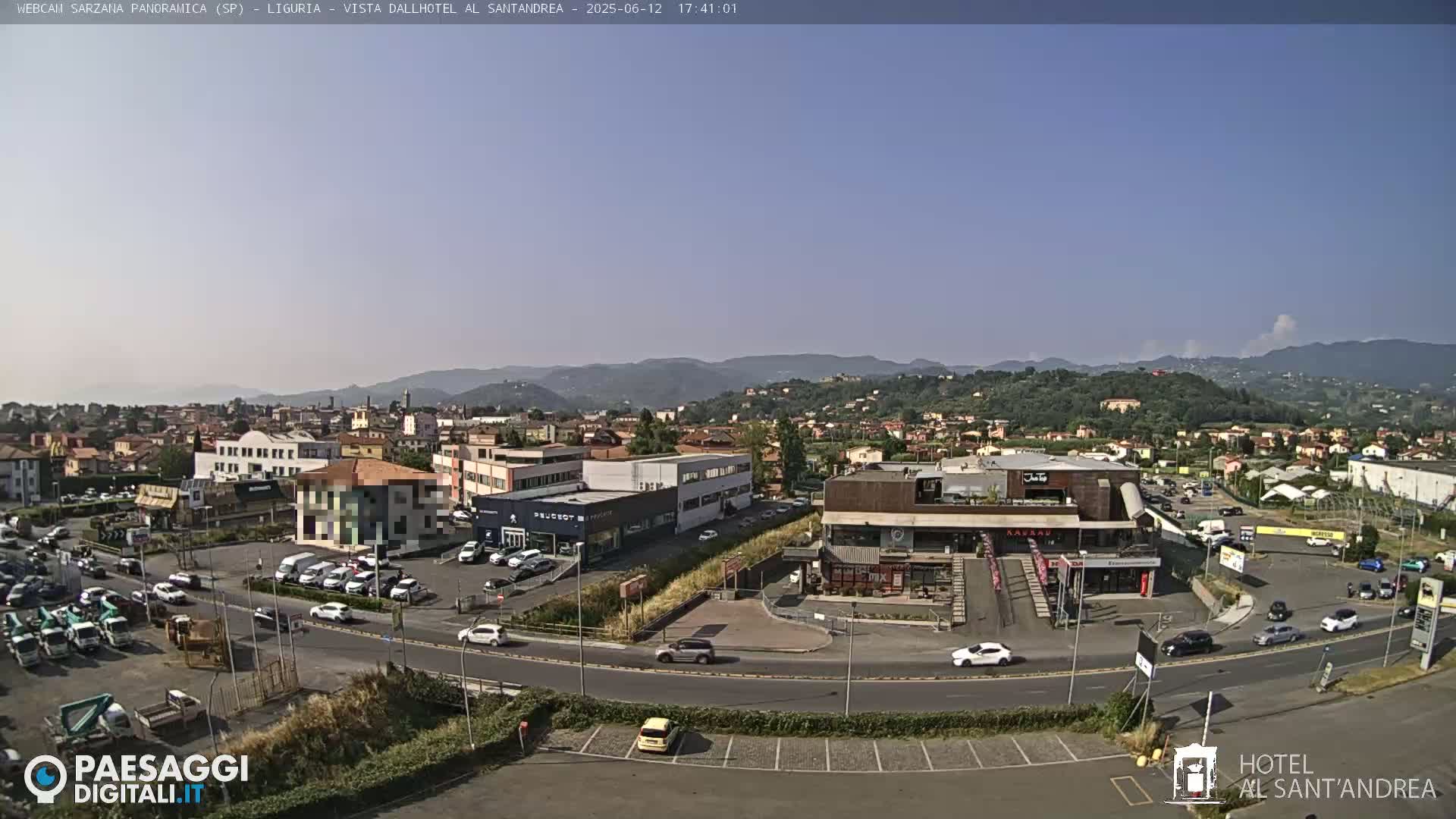 A sunny, clear day reveals a wide shot of a town with buildings, roads, and cars, nestled at the base of rolling hills.