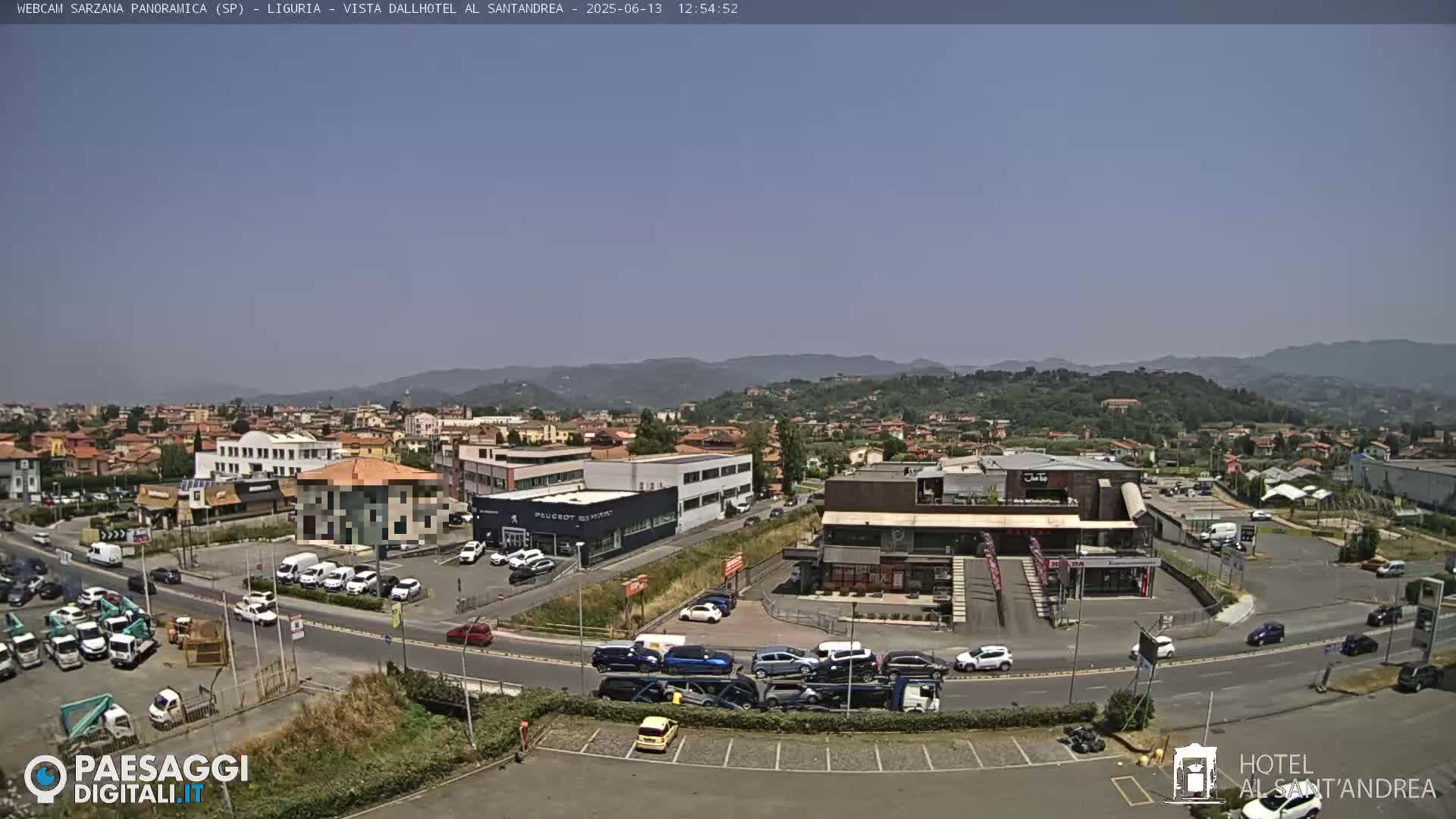 A sunny, clear-day view of a town situated in a valley, showing buildings, roads, vehicles, and distant hills.