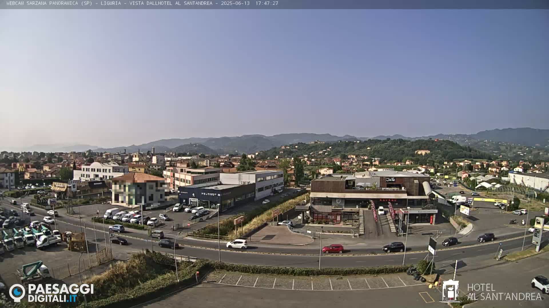 A clear, sunny day reveals a panoramic view of a town with buildings, cars, and roads, set against a backdrop of rolling hills.