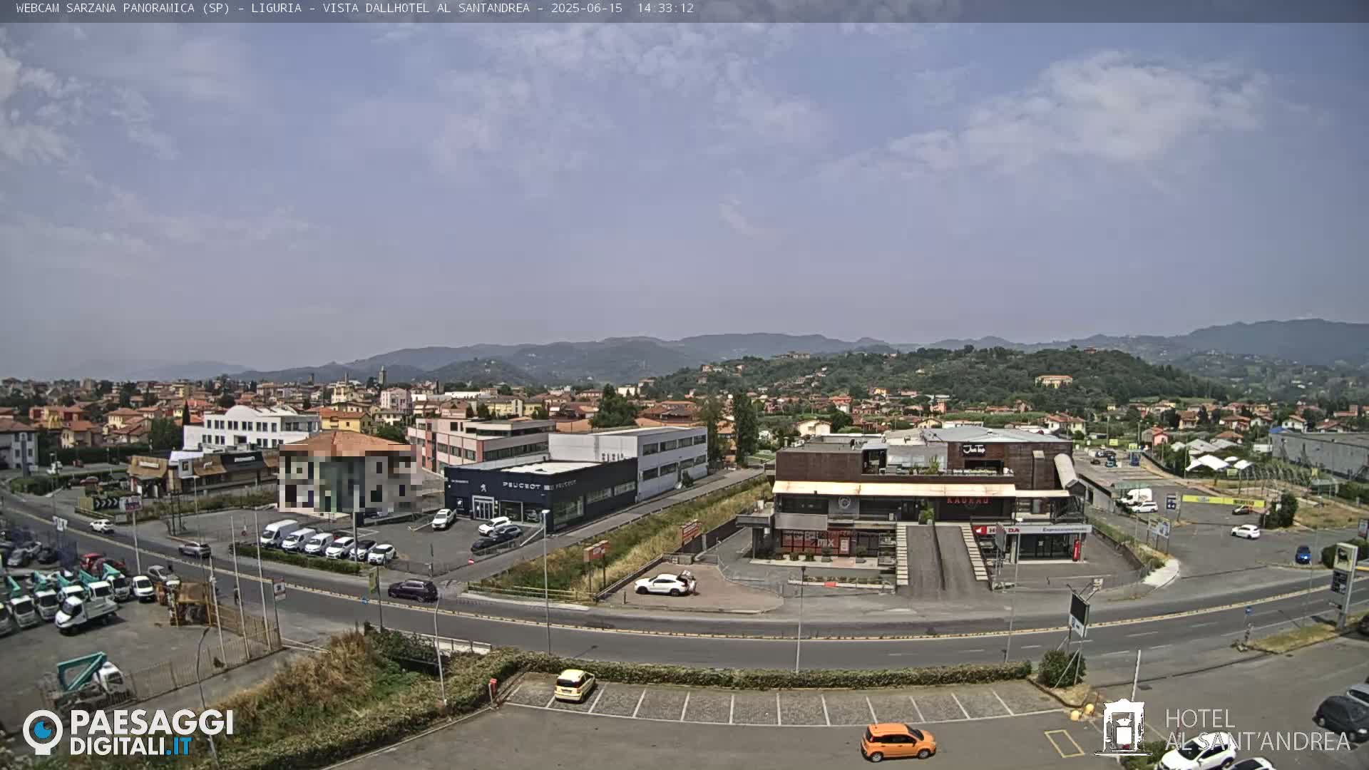 A mostly sunny, daytime view of a town nestled in a valley between hills, showing buildings, roads, and parked cars.