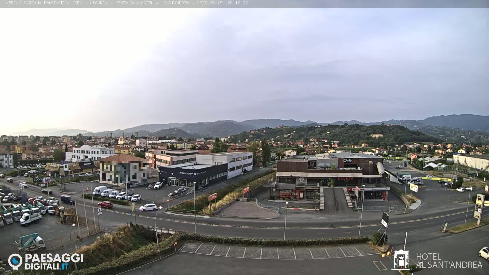 A wide, high-angle view shows a town nestled among hills under a mostly clear, light-grey sky, with cars parked in lots and driving on roads around various buildings.