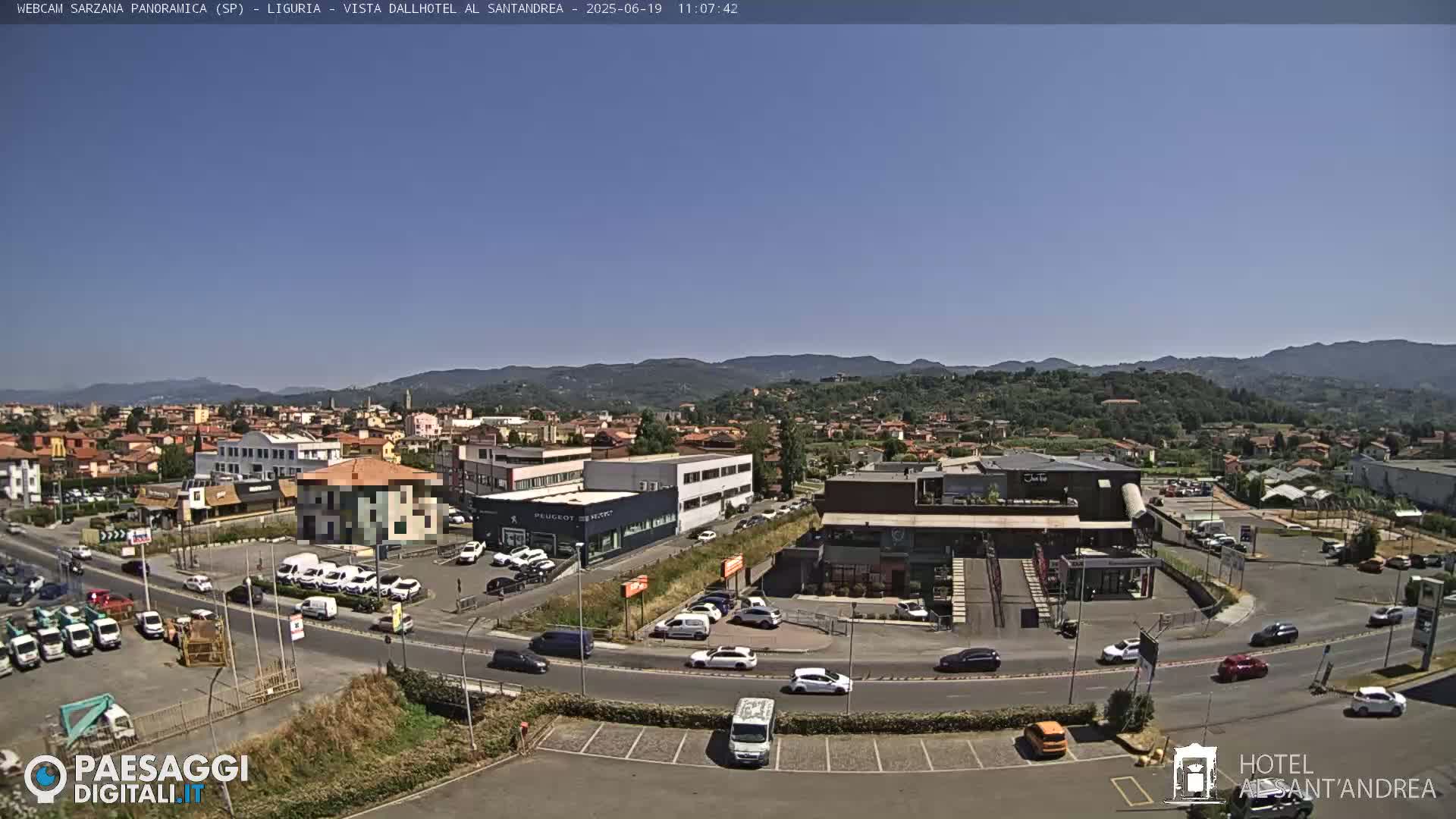 A clear, sunny day reveals a wide shot of a town nestled amongst green hills, with various buildings, cars, and a parking lot visible.