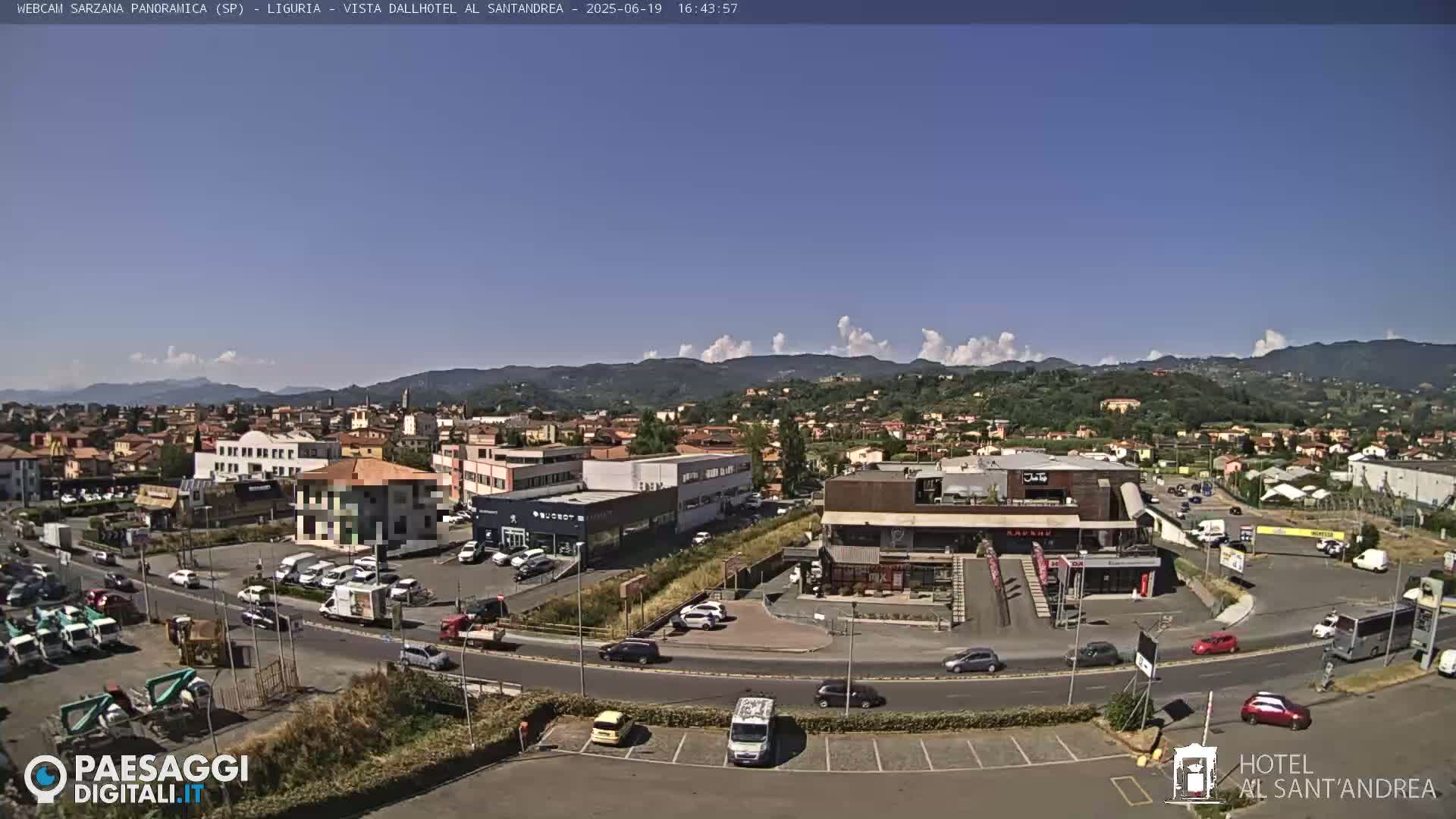 A sunny day reveals a town's cityscape, featuring buildings, roads with vehicles, and mountains in the background under a mostly clear sky with some cumulus clouds.