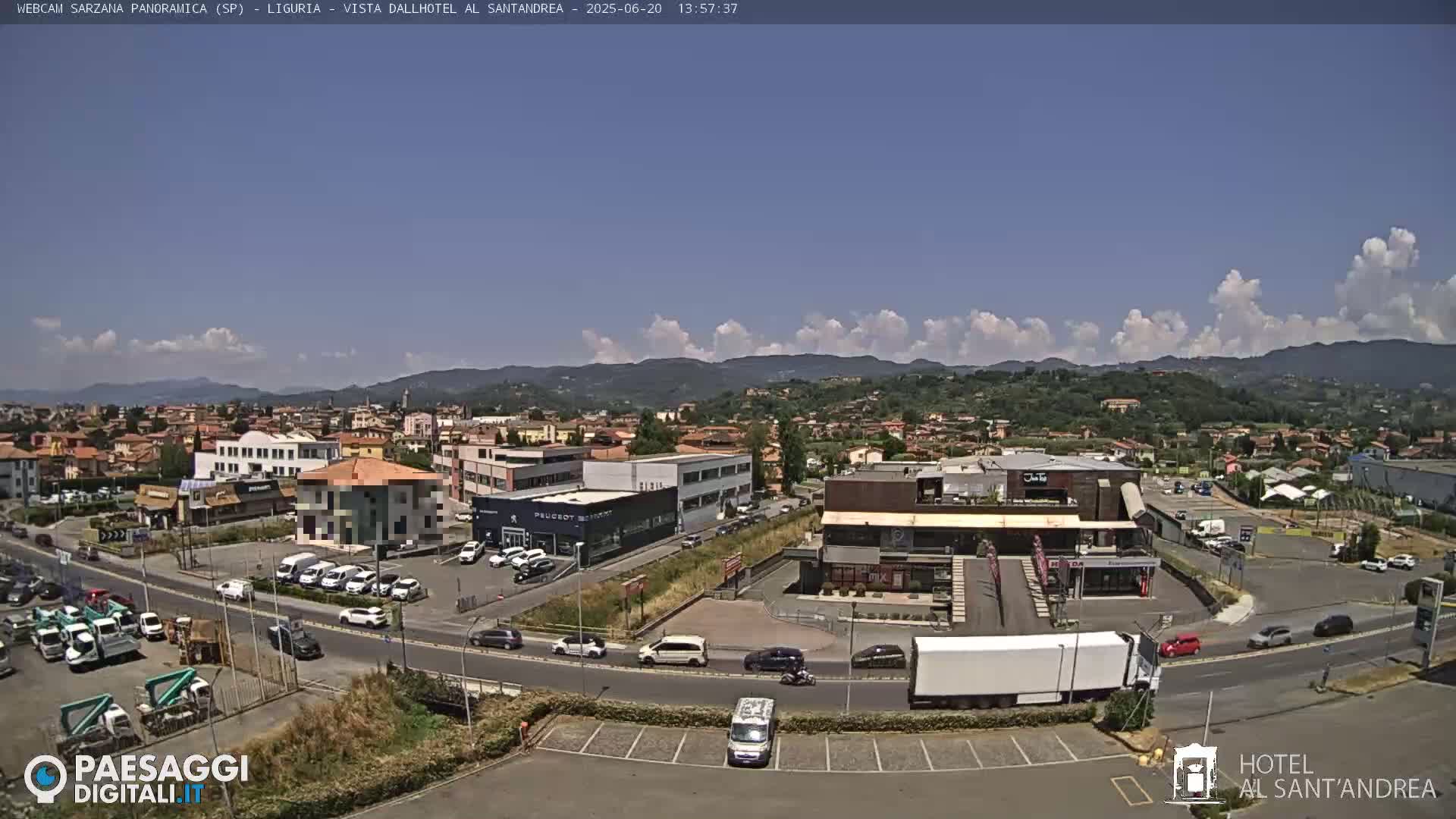 A sunny day reveals a panoramic view of a town nestled in a valley, surrounded by hills and mountains under a partly cloudy sky.