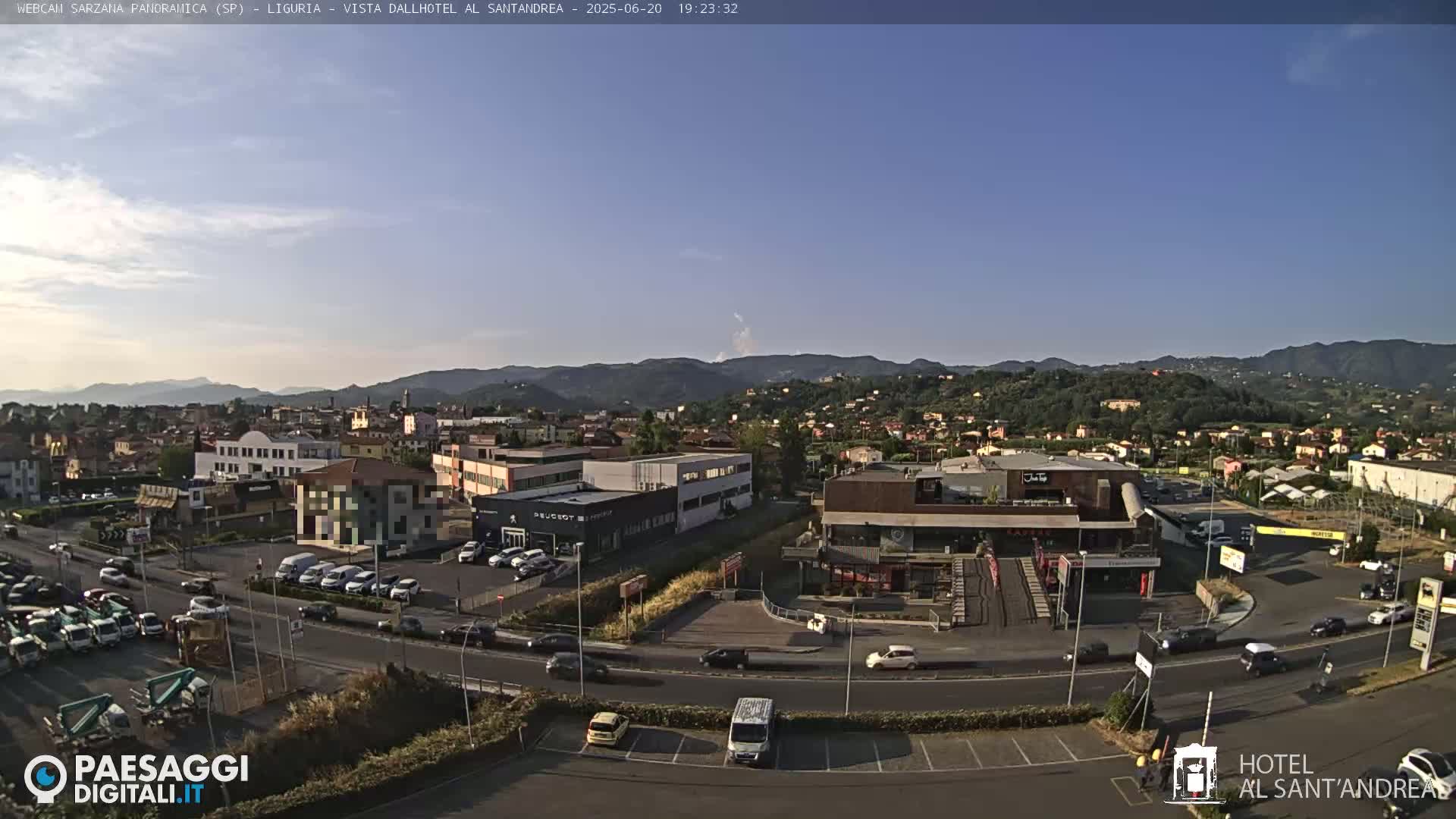 A clear, sunny day shows a panoramic view of a town nestled in a valley surrounded by hills and mountains, with cars parked in lots and driving on roads near various buildings, including car dealerships and restaurants.