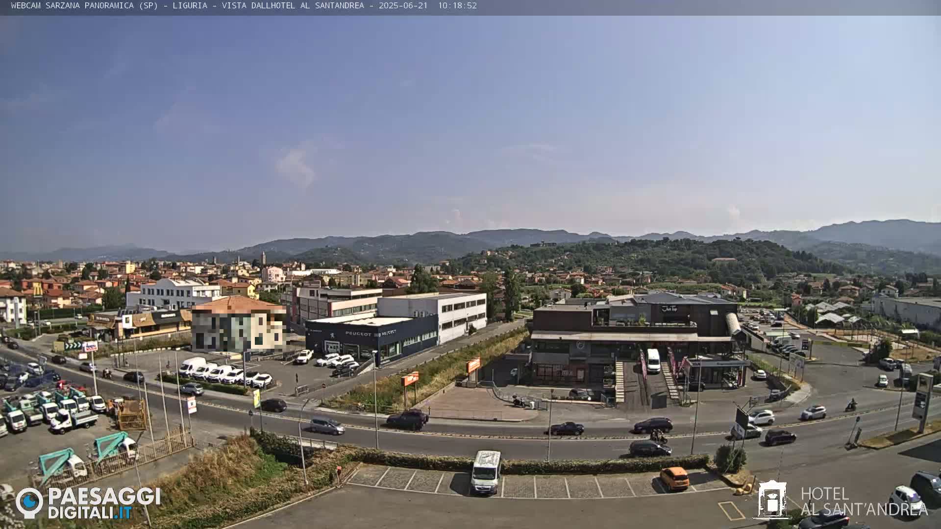 A sunny day reveals a wide shot of a town's outskirts, featuring buildings, roadways, parked cars, and distant hills under a clear sky.