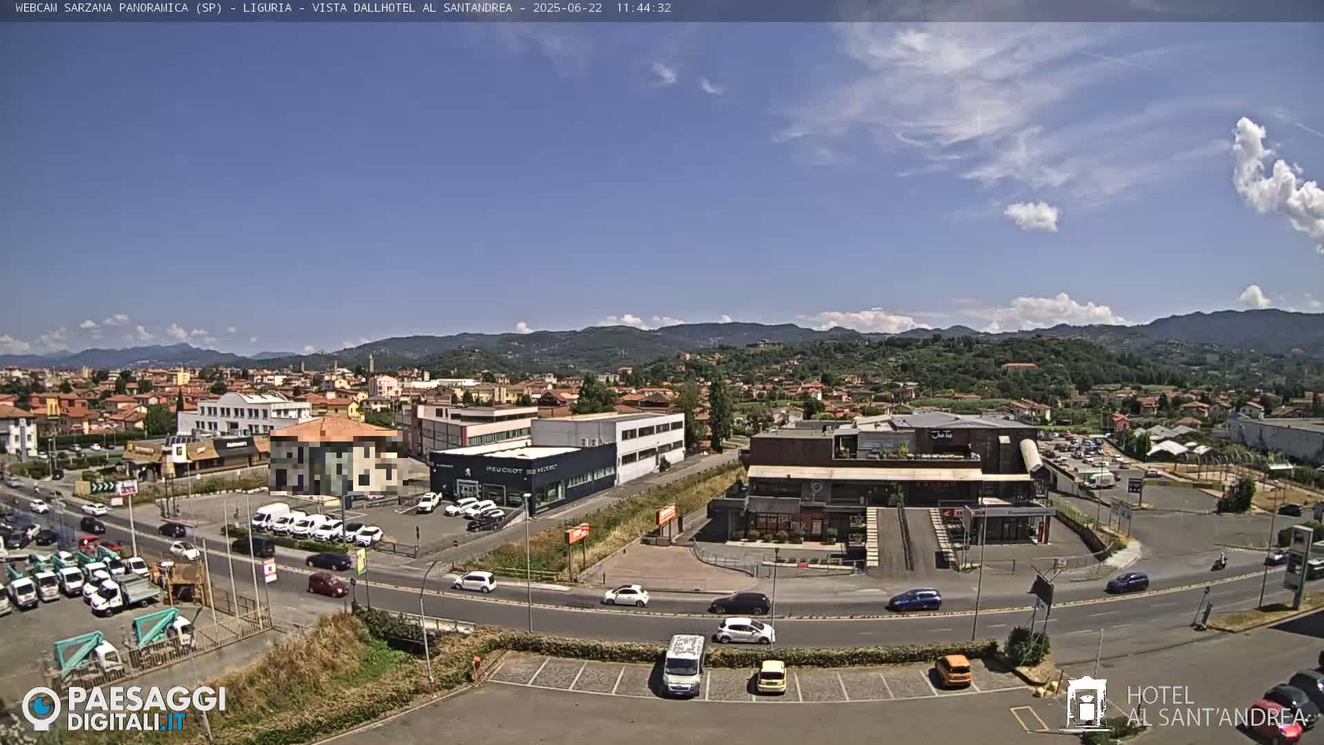 A sunny day reveals a panoramic view of a town nestled in a valley, with mountains in the background and various buildings, cars, and roads visible in the foreground.