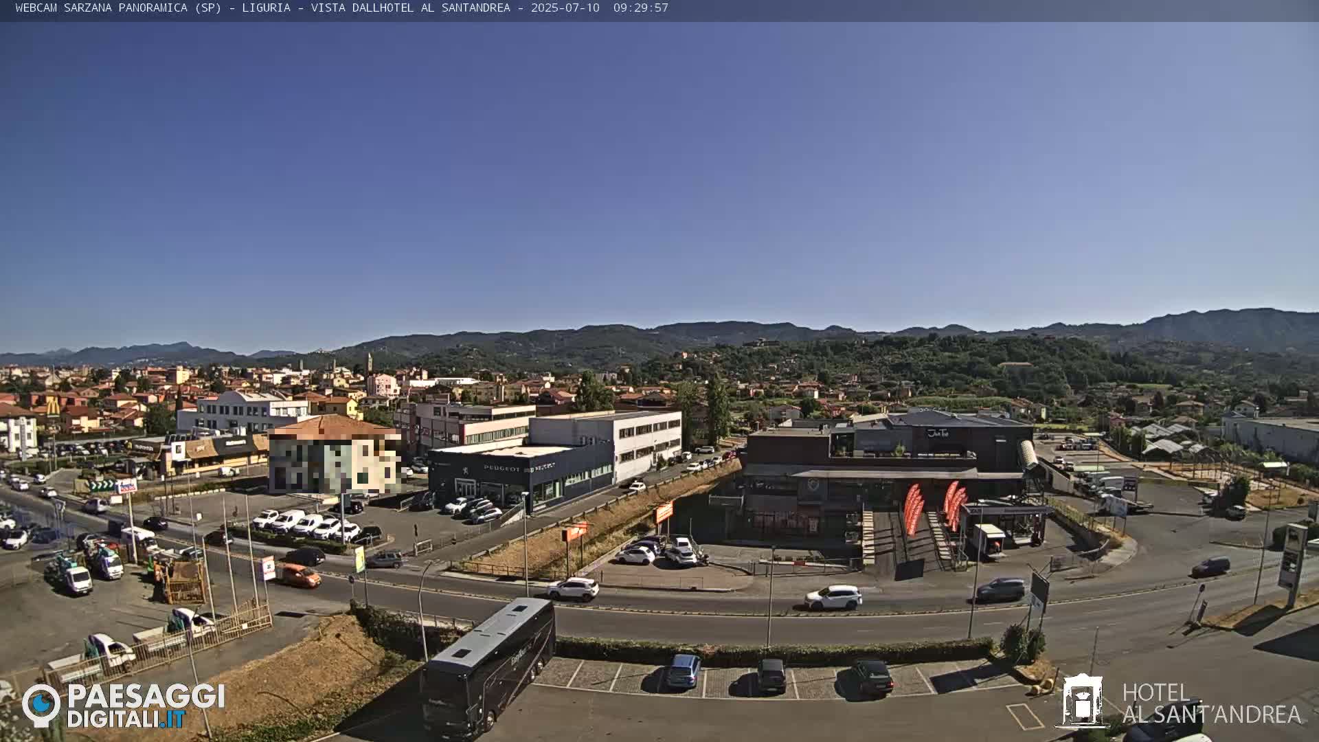 A sunny, clear day reveals a panoramic view of a town nestled in a valley, with mountains in the background, cars parked in lots, and a bus near a building.
