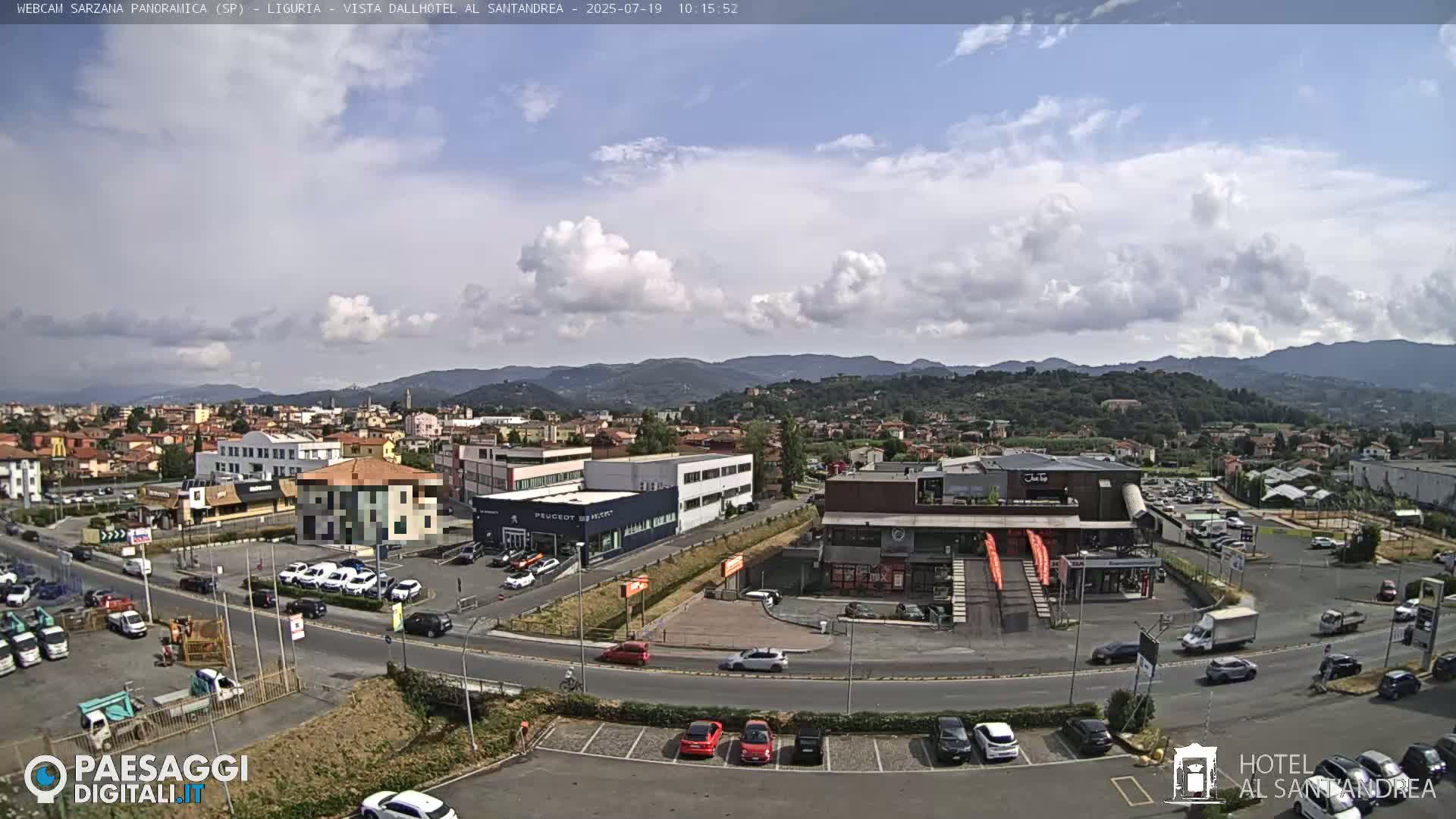 A partly cloudy day reveals a panoramic view of a town nestled among rolling hills, with cars parked in a lot near several buildings and a road curving through the scene.