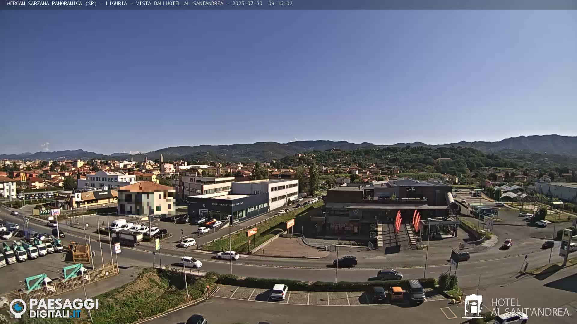 A clear, sunny day reveals a panoramic view of a town nestled in a valley, surrounded by mountains, with cars parked in lots and driving on roads around various buildings.