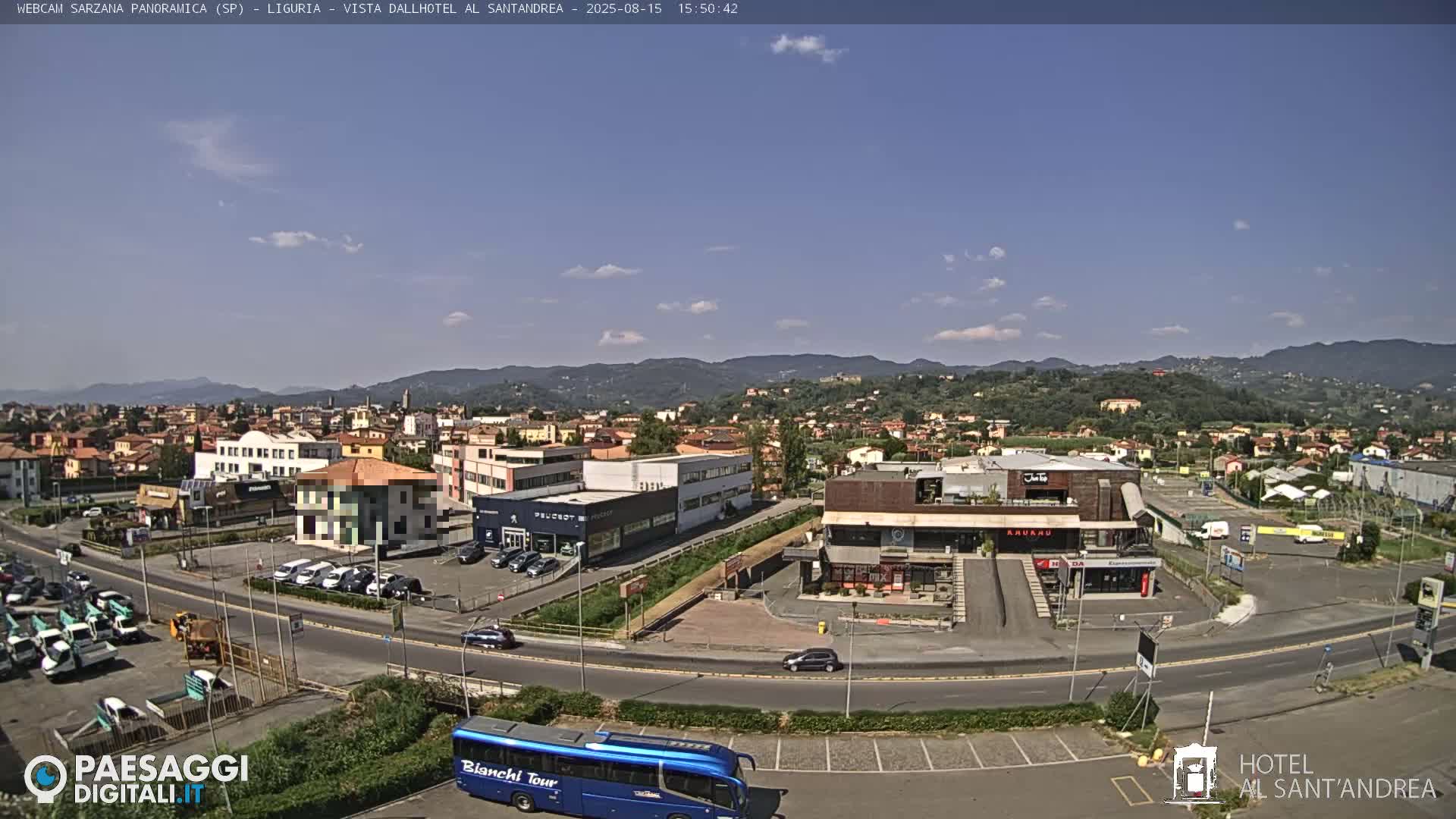 A clear, sunny day reveals a panoramic view of a town nestled in a valley, surrounded by mountains, with cars parked in lots and driving on roads around various buildings.