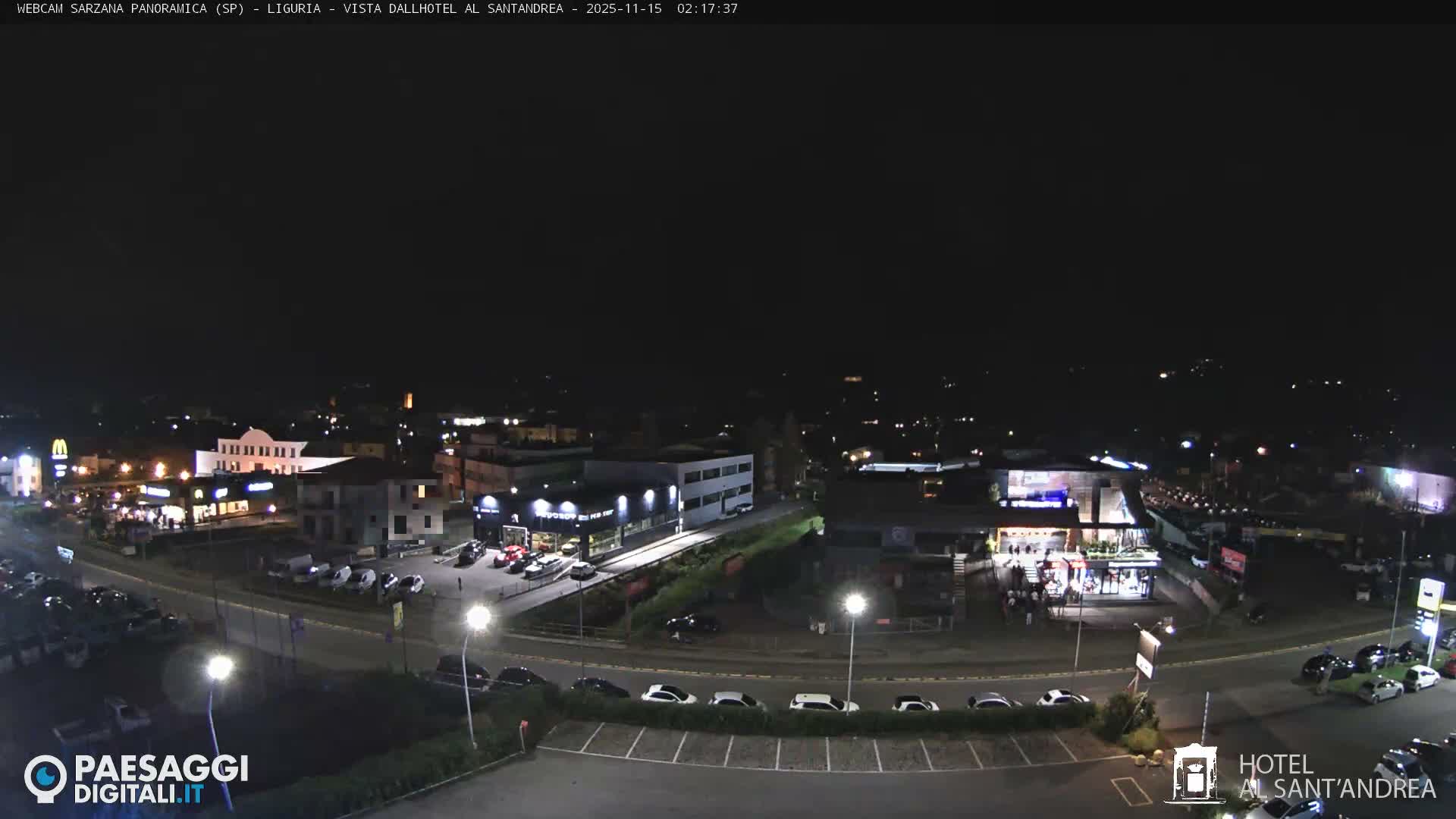 A nighttime panoramic view reveals an illuminated commercial area with buildings, roads, and parked cars, all visible under a clear, dark sky.