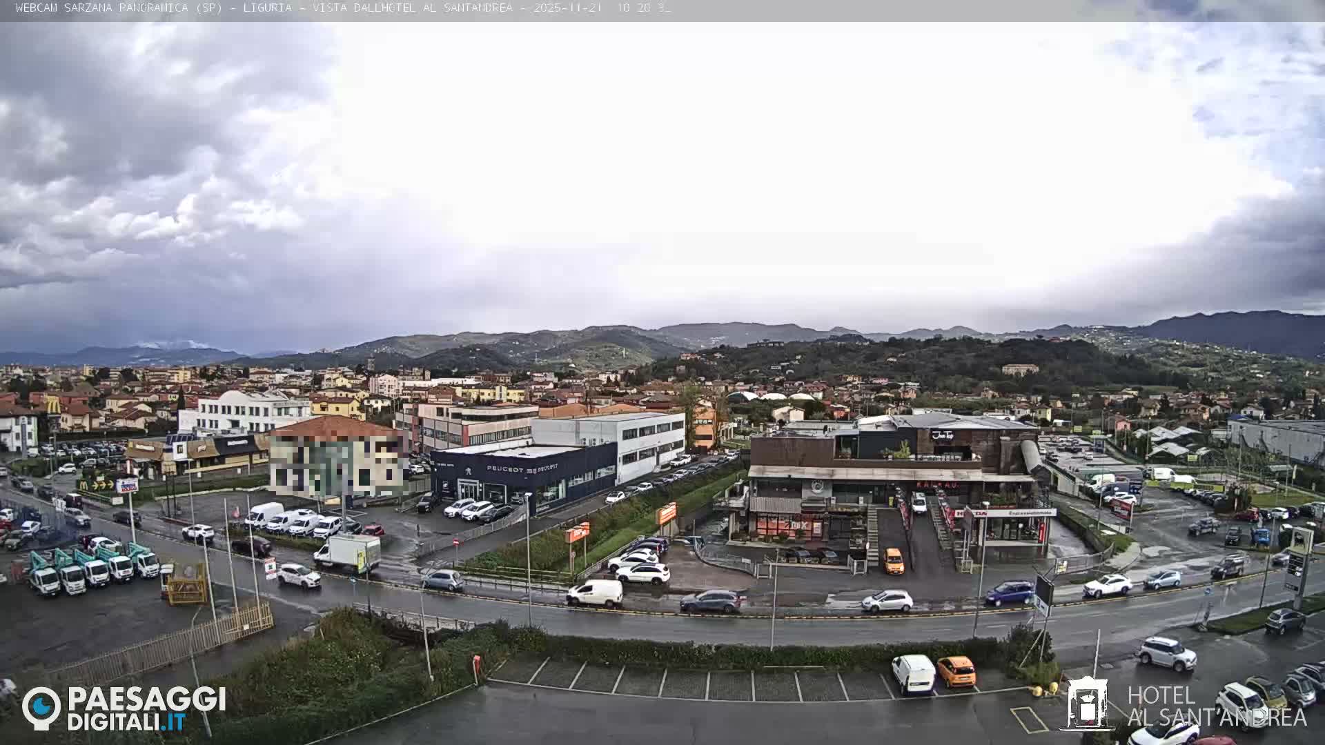 An elevated view captures a sprawling town with cars on wet streets and in parking lots, surrounded by commercial and residential buildings, all set against a backdrop of green, undulating hills and distant mountains under a dark, heavily overcast sky.