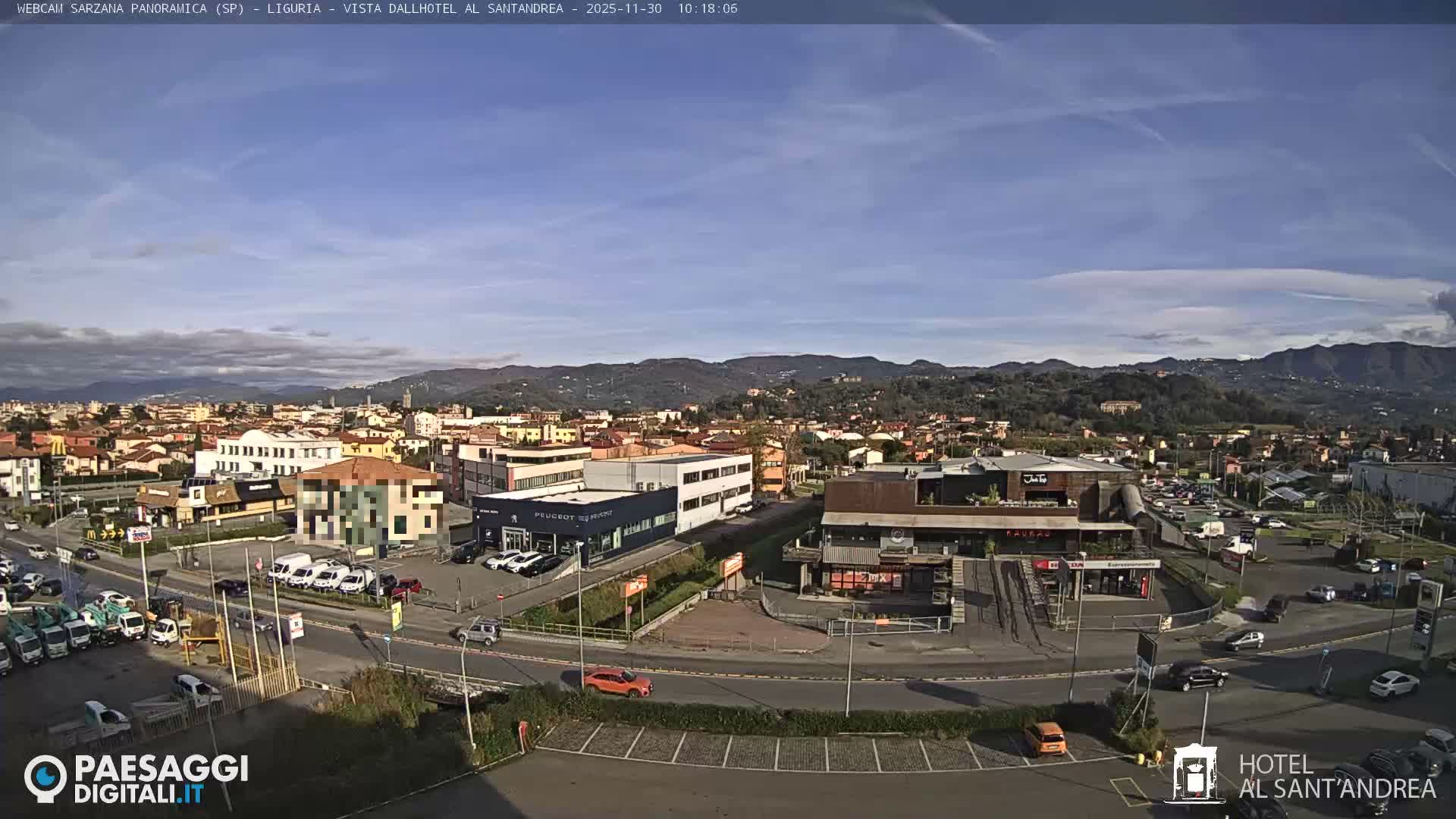 A wide-angle daytime view captures a bustling urban area with roads, numerous cars, and various buildings, all framed by distant green, tree-covered hills under a bright, partly cloudy sky.