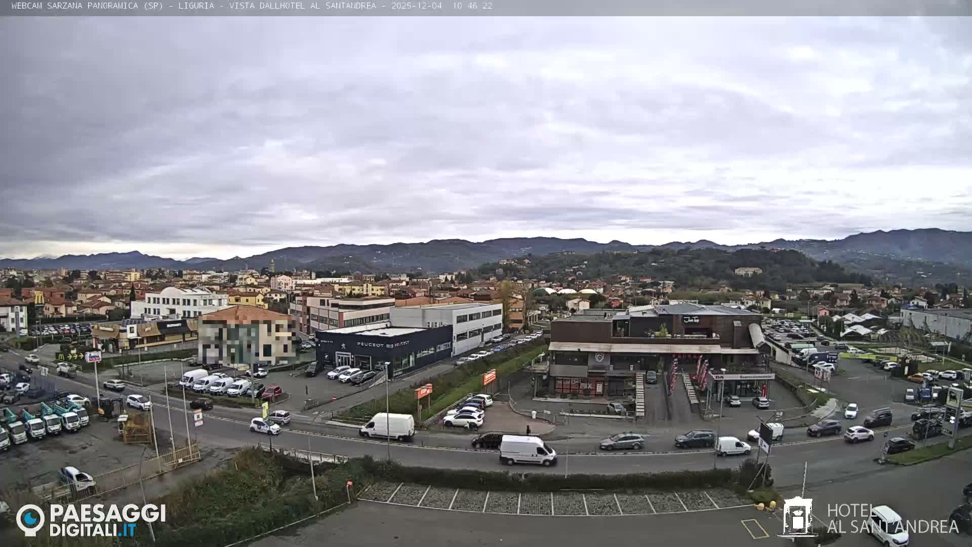 An expansive overhead view reveals a moderately busy town situated at the base of lush green hills under a uniformly overcast sky, with commercial buildings, residences, and numerous cars populating its streets and parking areas.
