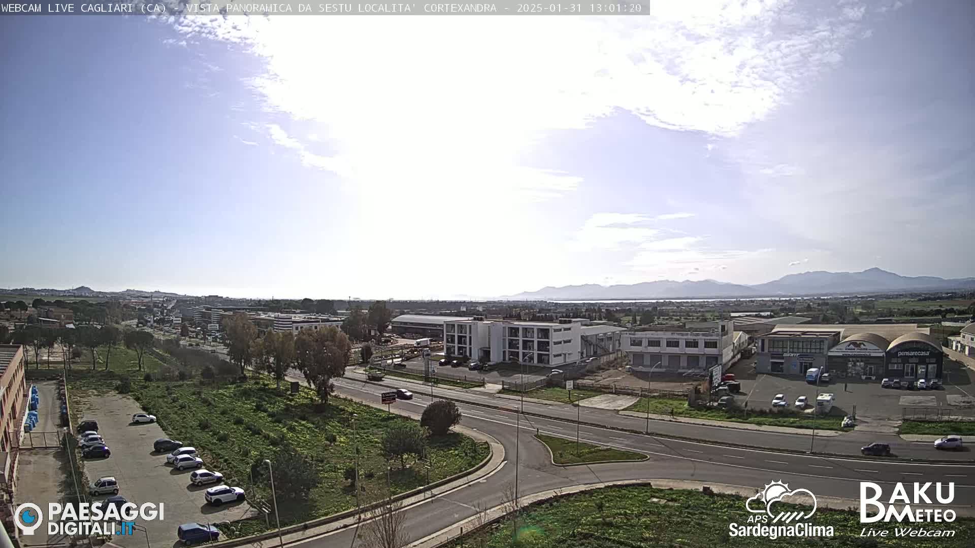 A panoramic view of a city with clear skies and distant mountains, showing a road intersection with several buildings and parked cars.