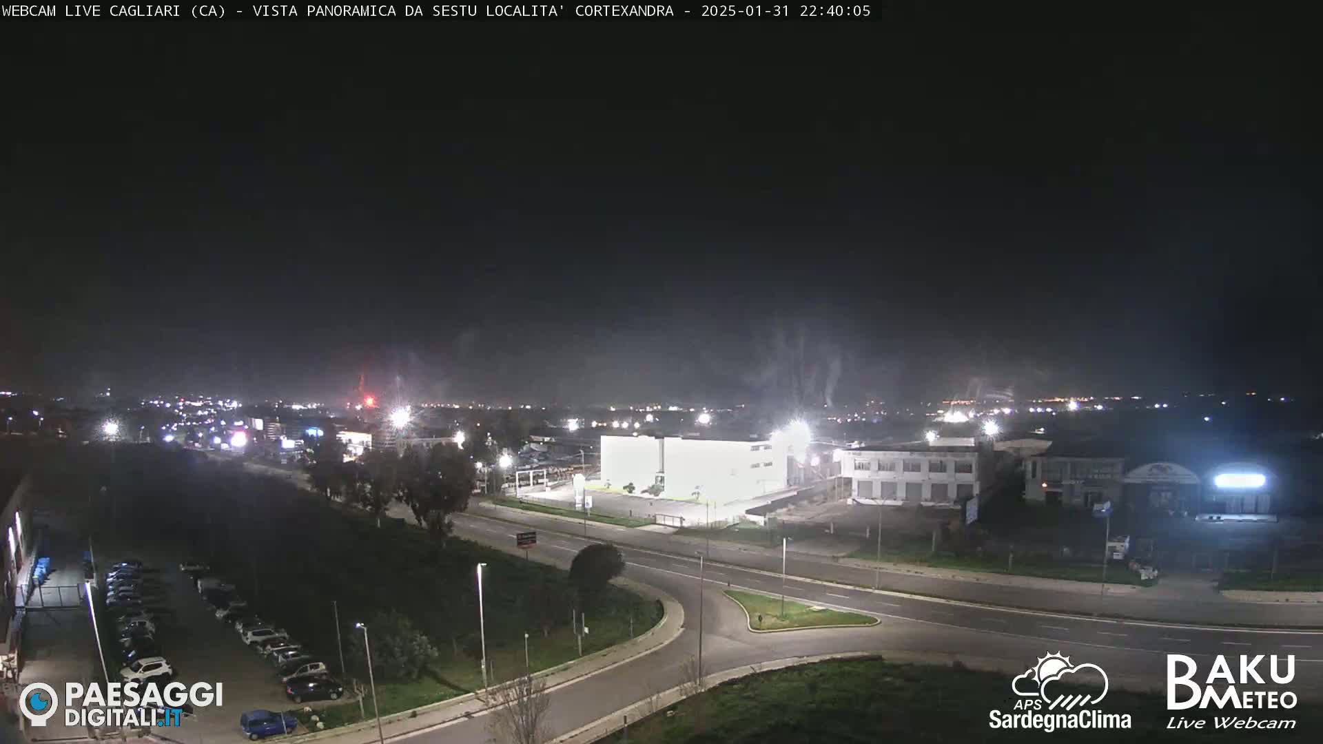 A nighttime, aerial view of a road intersection and industrial area, with some light haze or smoke in the air.