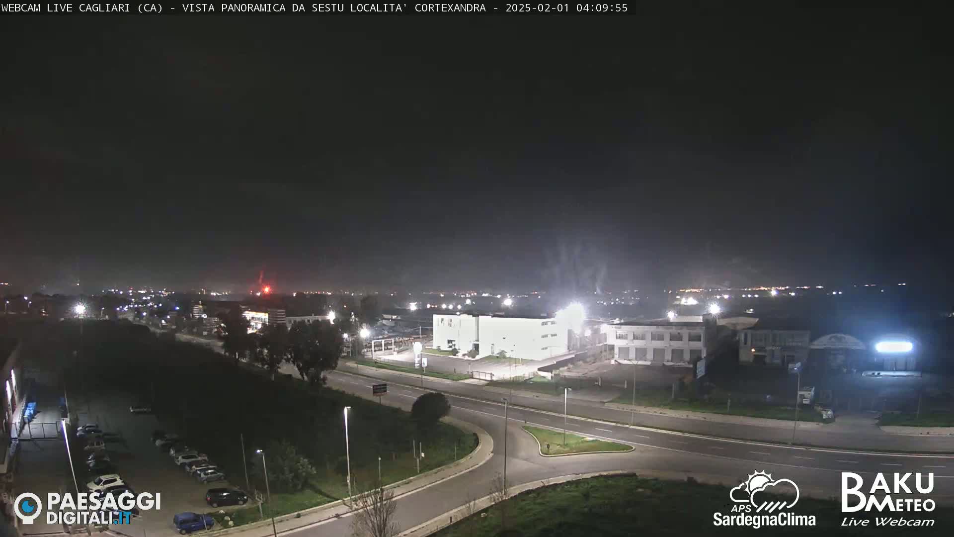 A nighttime aerial view of a road intersection and industrial area, with a dark sky and faint lights in the distance.