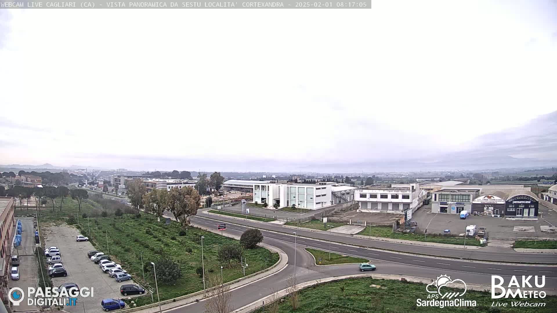 A mostly cloudy day reveals a wide view of an industrial area and parking lot next to a road intersection, with a distant cityscape visible under a hazy sky.