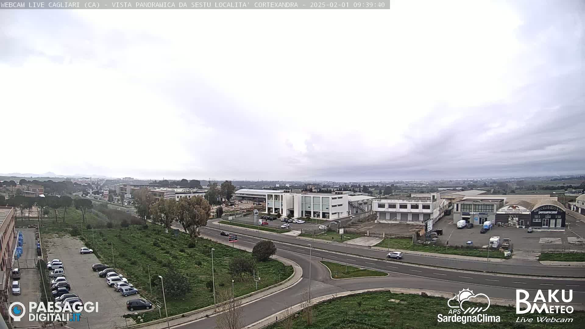 A panoramic view of a city under an overcast sky shows a highway intersection surrounded by industrial buildings, parking lots full of cars, and green spaces.