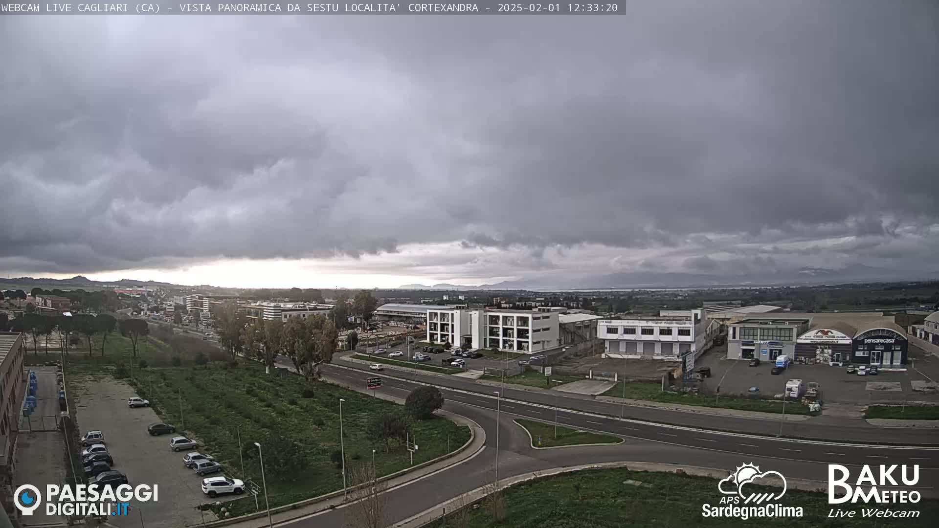 A panoramic view of a city with overcast skies shows a road intersection and various buildings, including industrial structures and apartment complexes, with some cars parked in a lot.