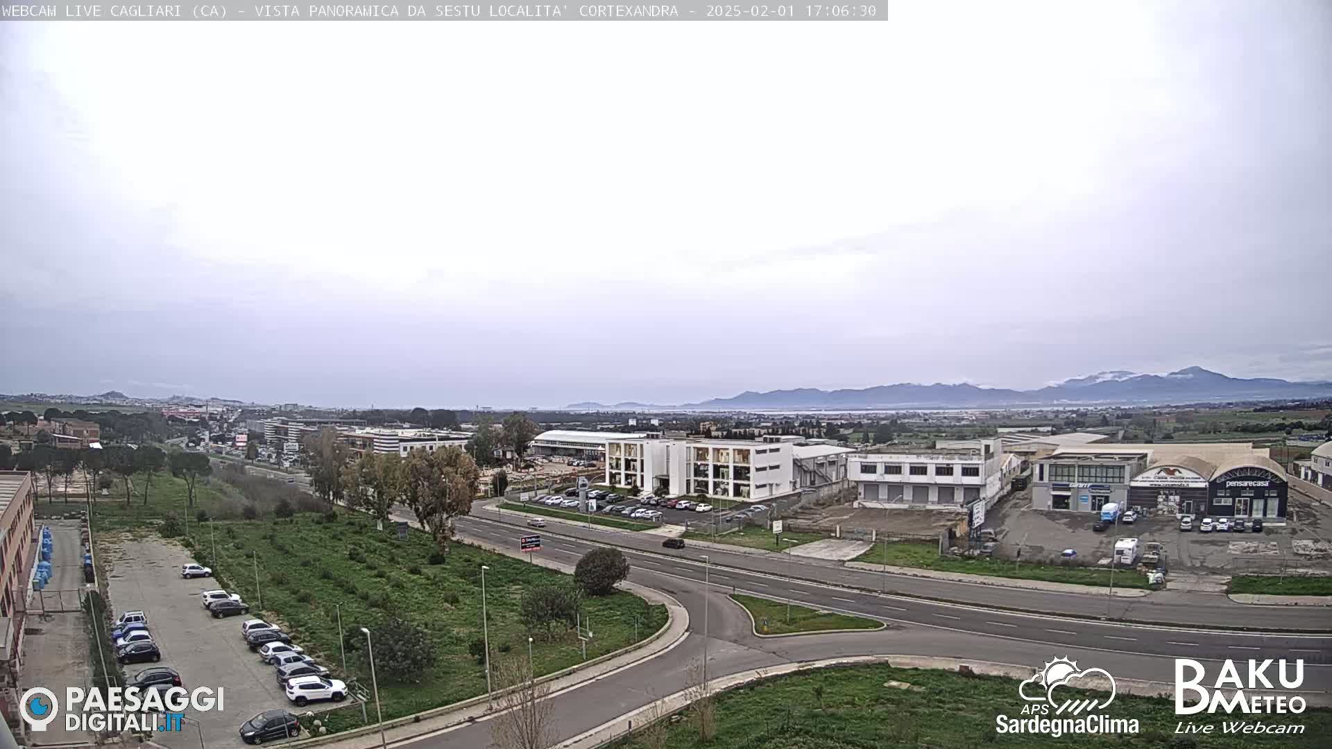 A panoramic view of a city with overcast skies shows a road intersection, parking lots filled with cars, and industrial buildings, with mountains visible in the distance.