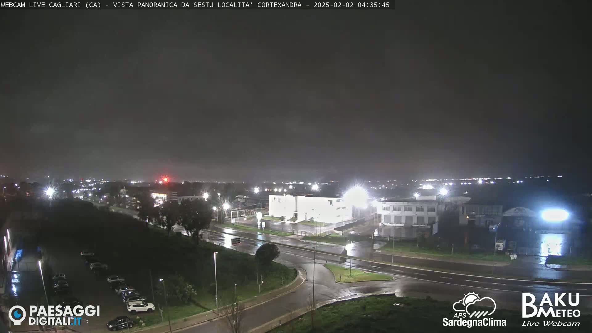 A nighttime view of a city intersection with wet roads under a dark, overcast sky.