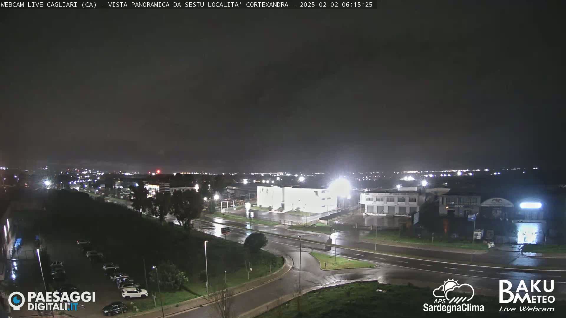 A nighttime, rain-slicked road intersection near a brightly lit industrial area, with a darker cityscape visible in the distance under a cloudy sky.