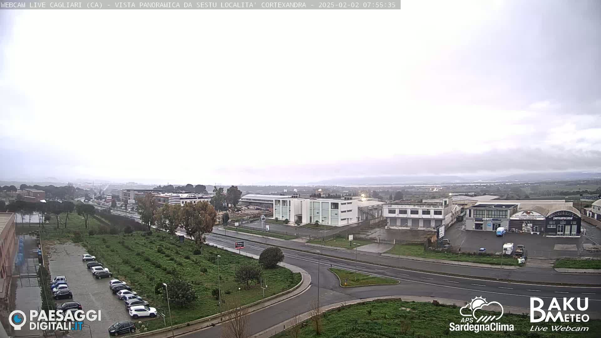 A wide shot of a city landscape on an overcast day, showing roads, buildings, and a parking lot filled with cars, with a body of water visible in the distance.