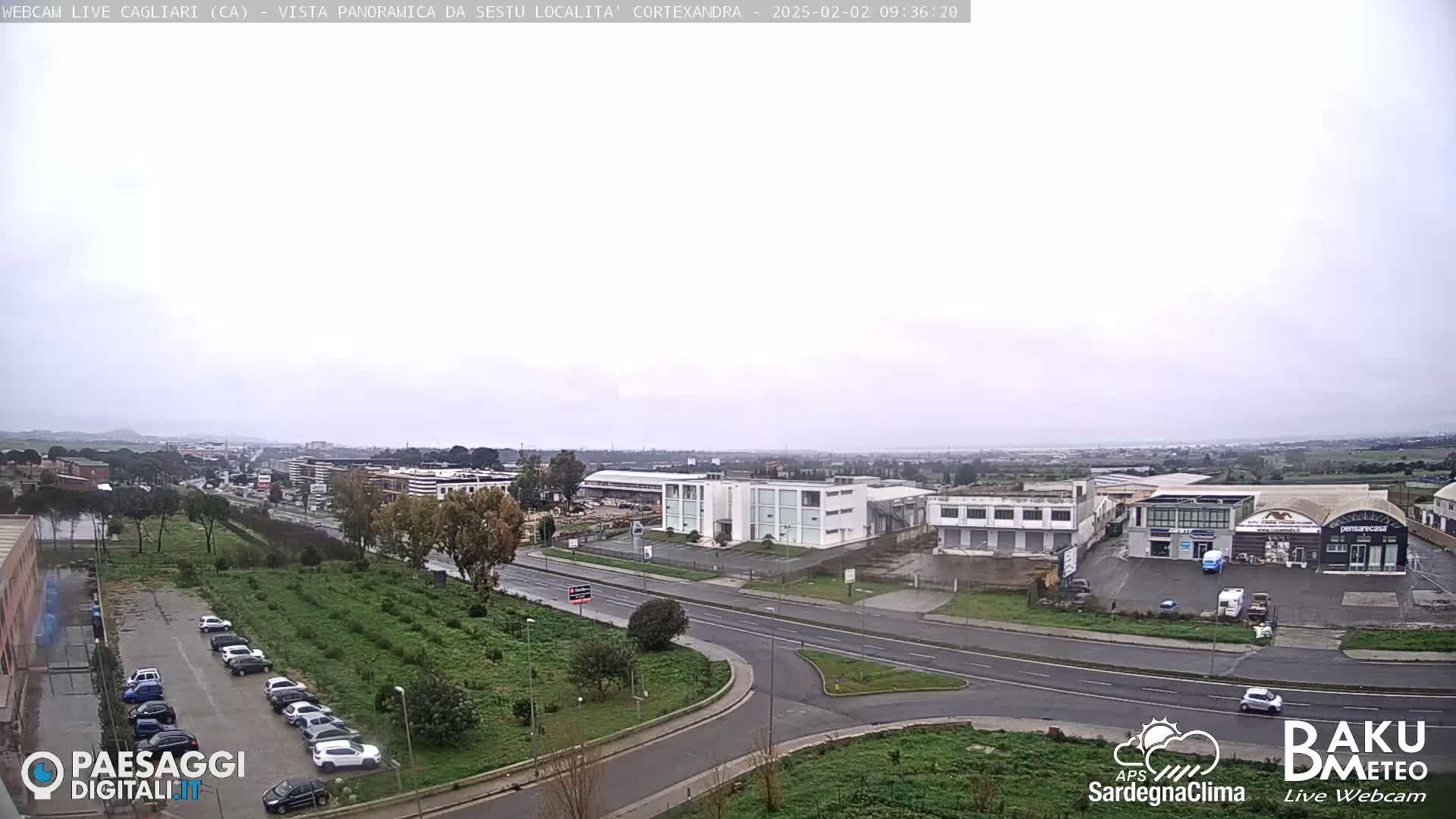 A wide, overcast view of a roadway and industrial area, with several cars parked in a lot and others traveling on the road.