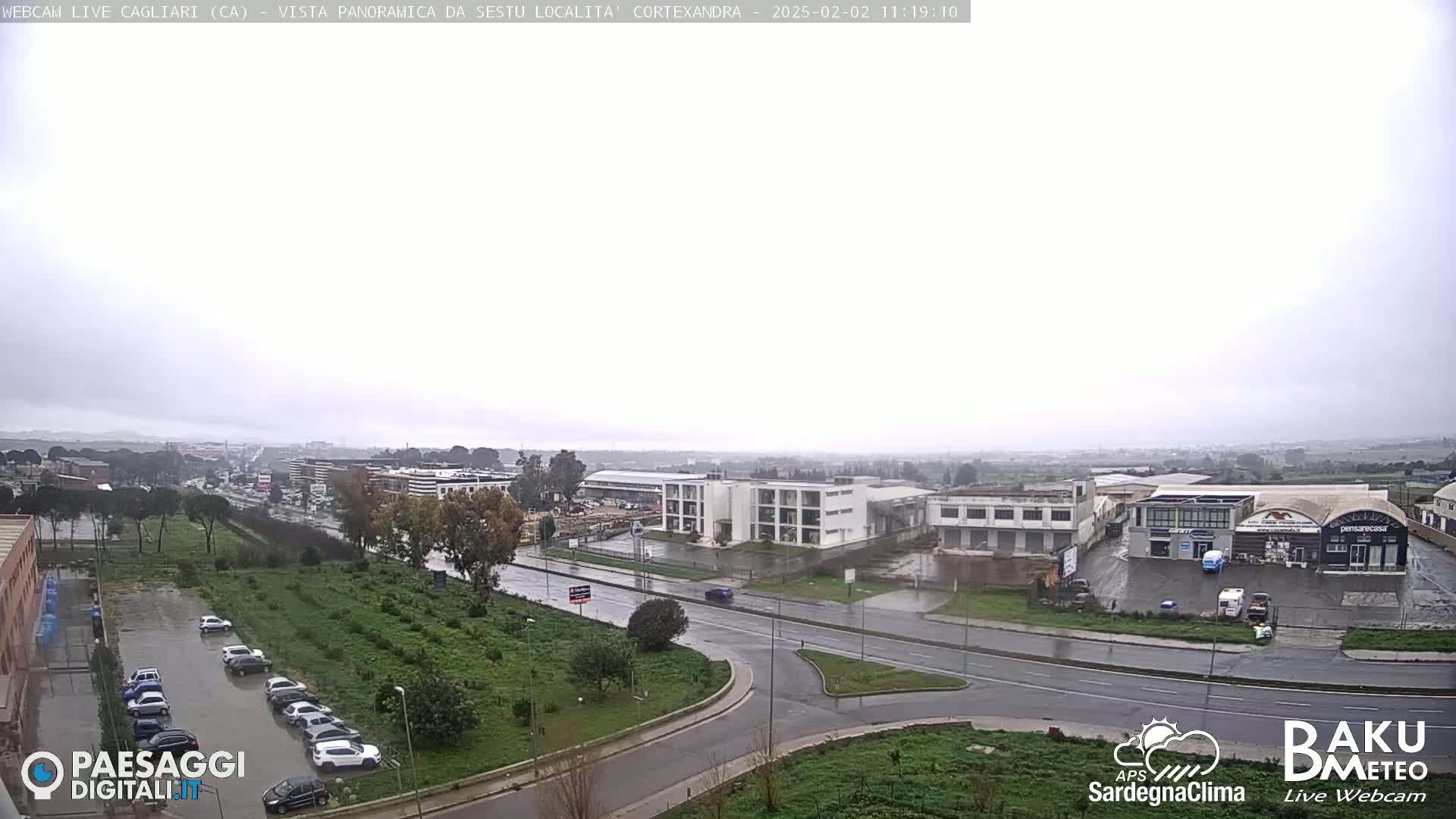 A wide, overcast view of a city showing roads, buildings, and parked cars in the rain.