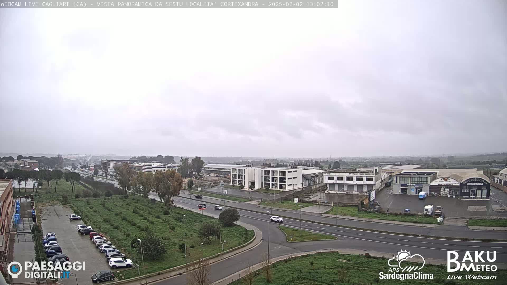 A wide, overcast view of a road intersection with buildings and a parking lot, surrounded by low vegetation.