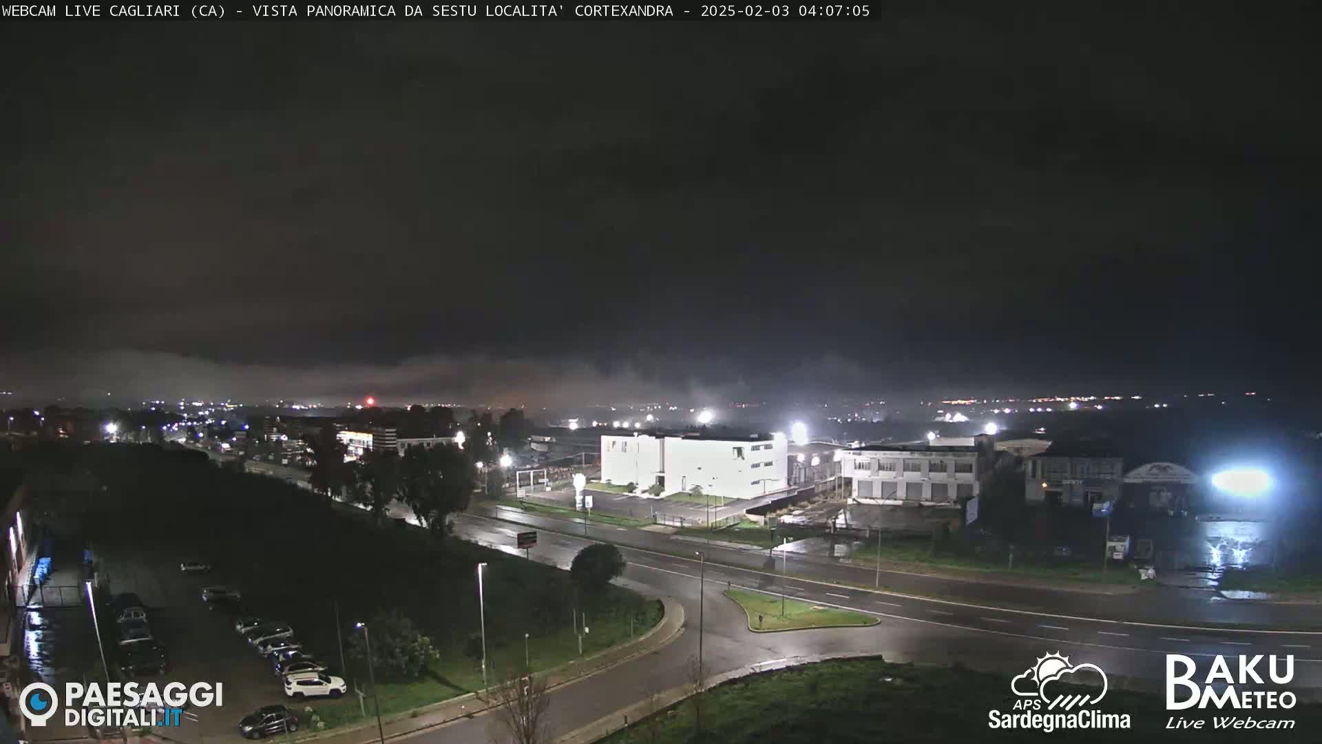 A nighttime, low-foggy view of a city road intersection and industrial area with some buildings illuminated under a dark sky.