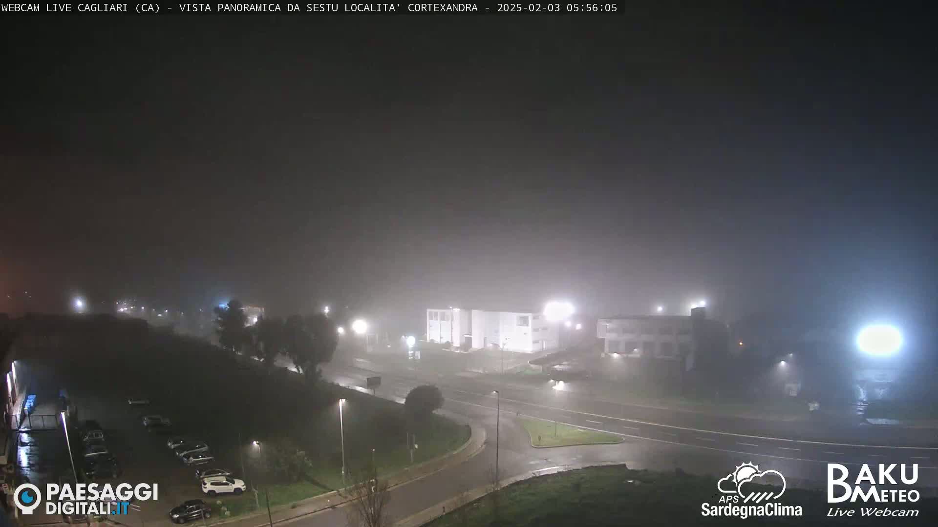 A nighttime, foggy scene shows a road intersection with cars parked in a lot and buildings in the background under dim lighting.