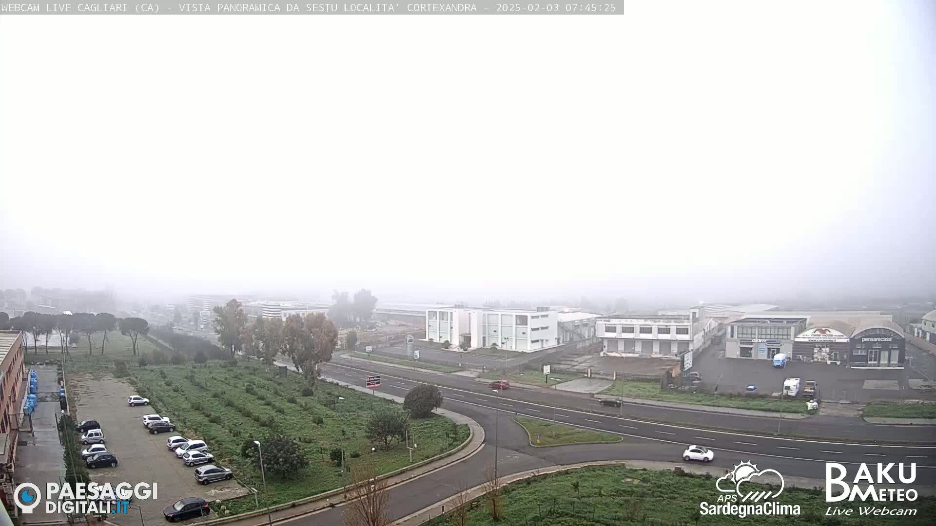 A foggy, overcast view of a road intersection surrounded by industrial buildings, a parking lot with cars, and green vegetation.