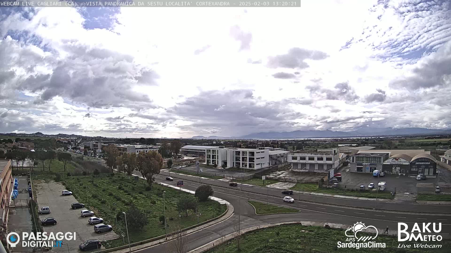 A mostly cloudy day reveals a wide view of a city with industrial buildings and roads, featuring distant mountains and a body of water under a partially clouded sky.